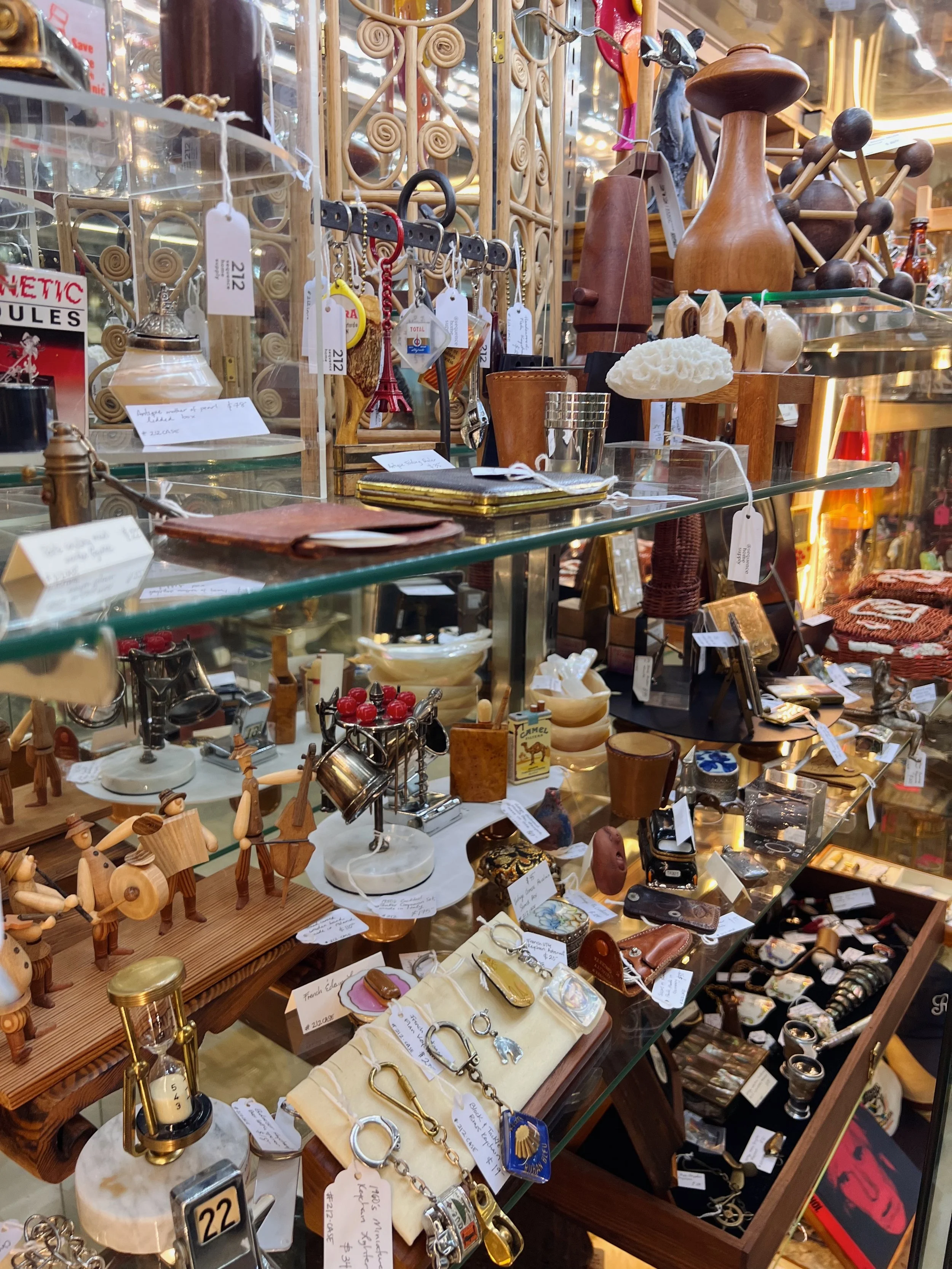 A display of various decorative and vintage items including wooden figurines, jewelry, Small metal objects, vases, and a mimicking lamp, all arranged on glass and wooden shelves in an antique shop.