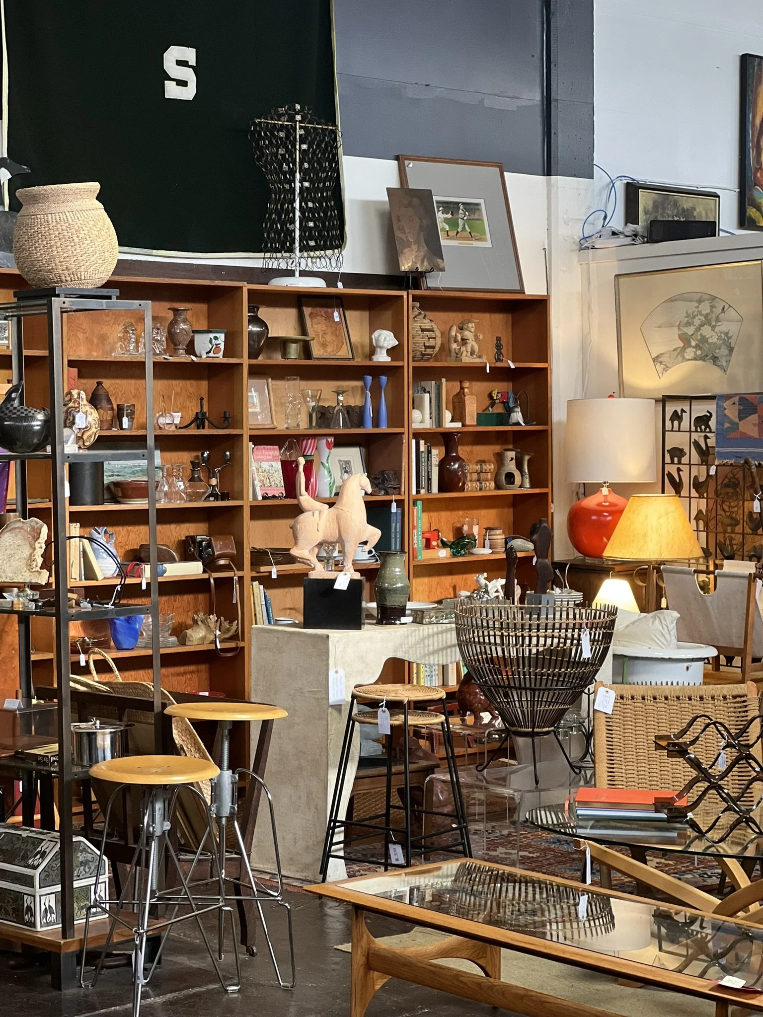 Interior of an antique shop featuring wooden shelves filled with various decorative items, picture frames, and sculptures, tables with glass tops, chairs, lamps, and artwork on the walls.