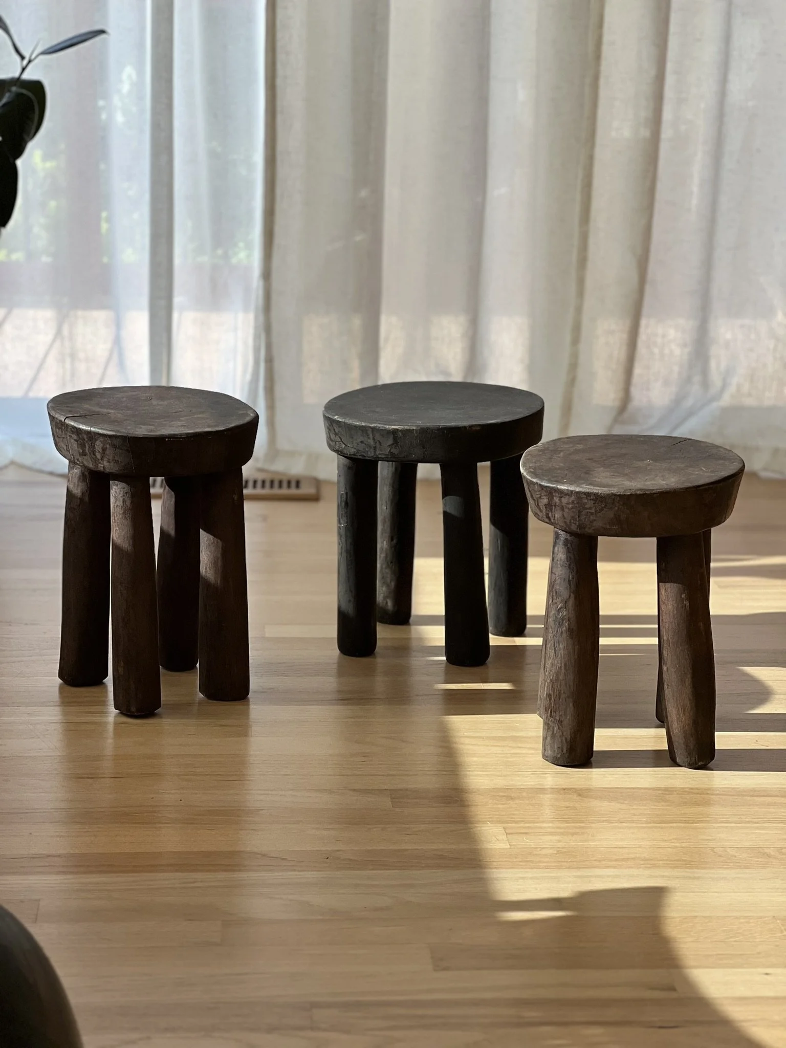 Four small wooden stools arranged in a row on a light wooden floor with a sheer curtain and sliding glass door in the background.