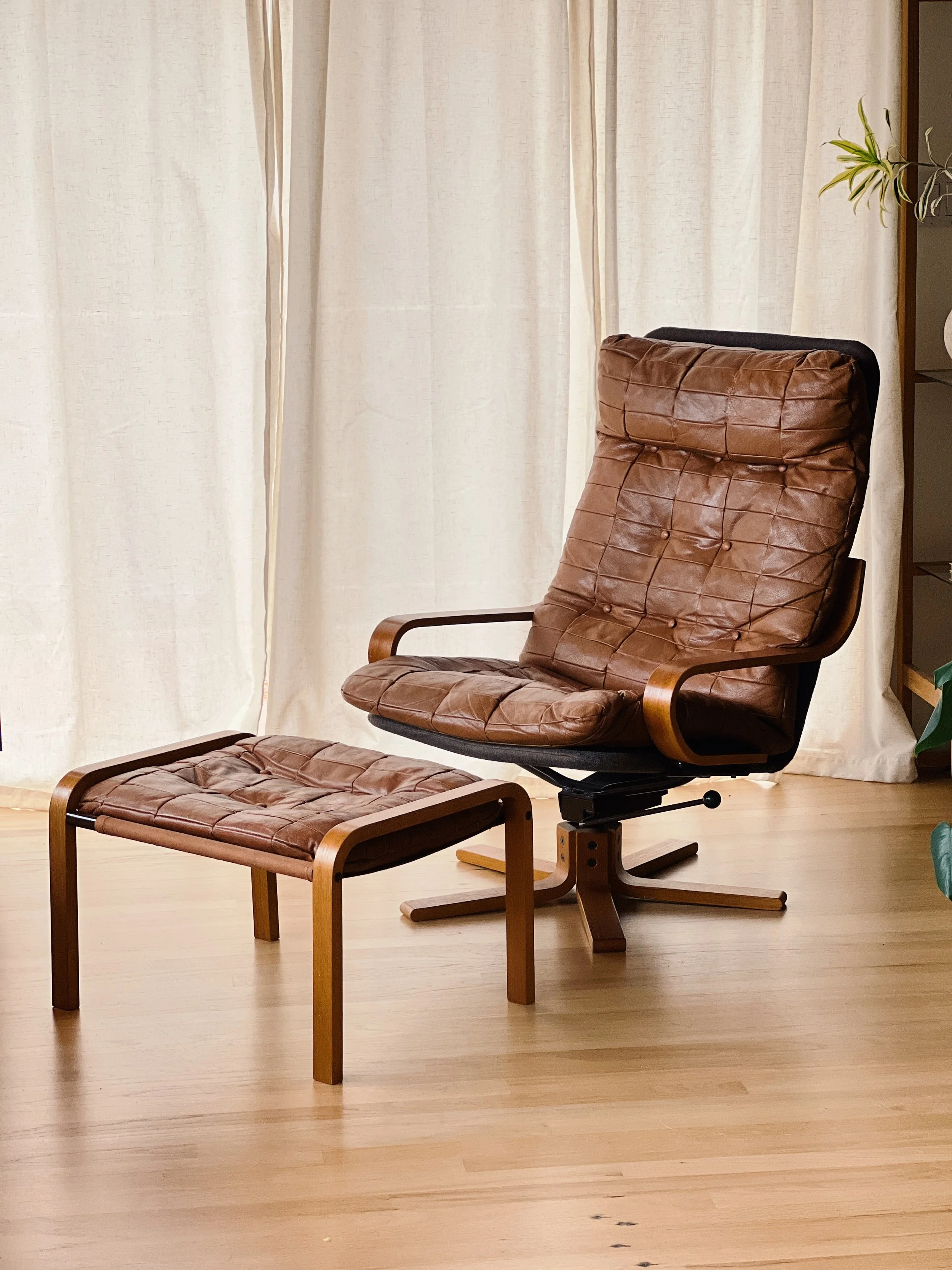A brown leather and wood armchair with matching footrest in front of cream curtains, on a light wood floor.