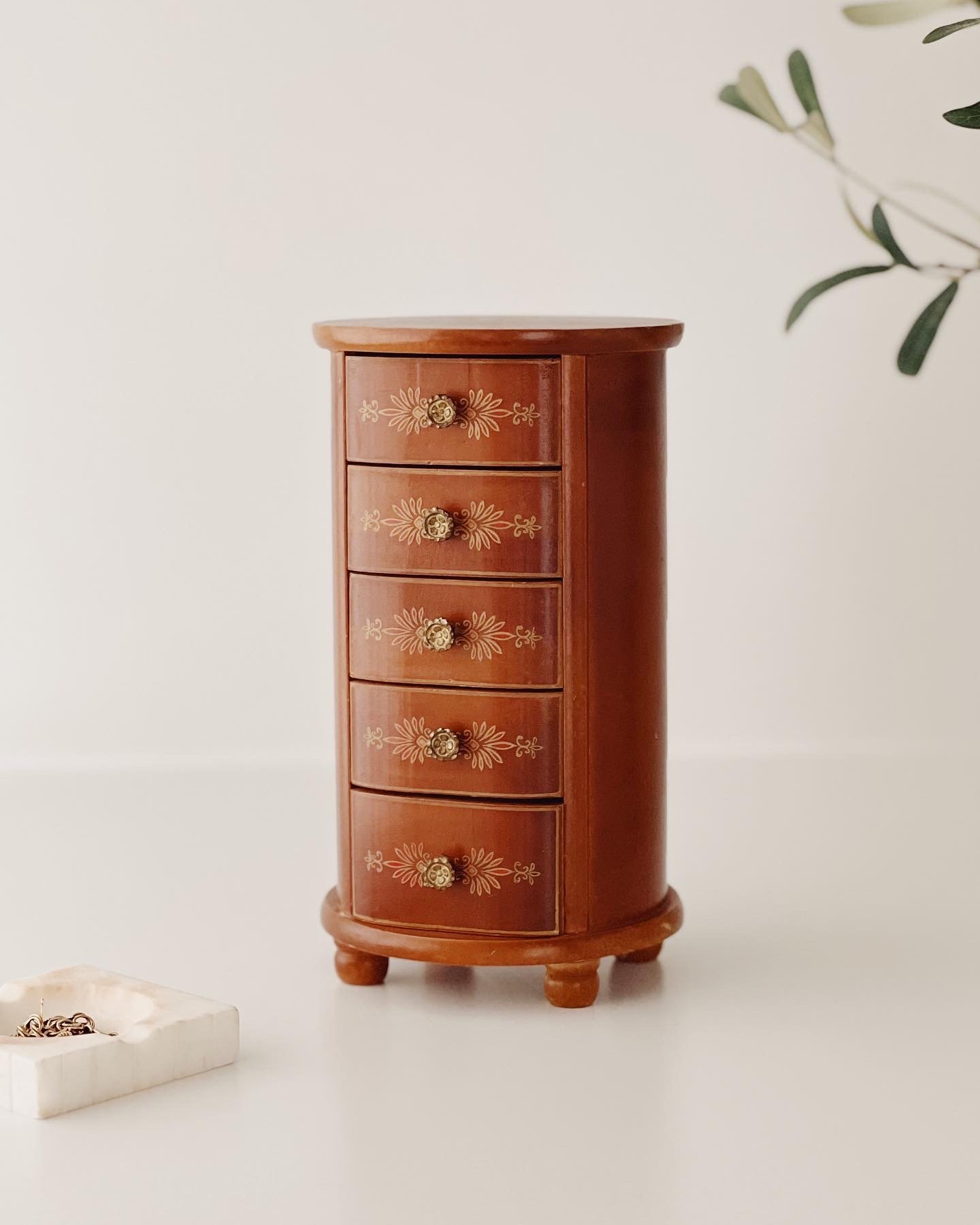 A small wooden jewelry cabinet with five drawers, decorated with gold floral patterns and round gold knobs, placed against a plain white background.