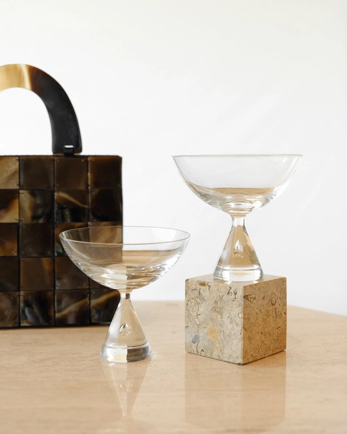 Two clear glass bowls with stems, one on a stone block and one on a light wooden surface, with a black and gold patterned bag in the background.