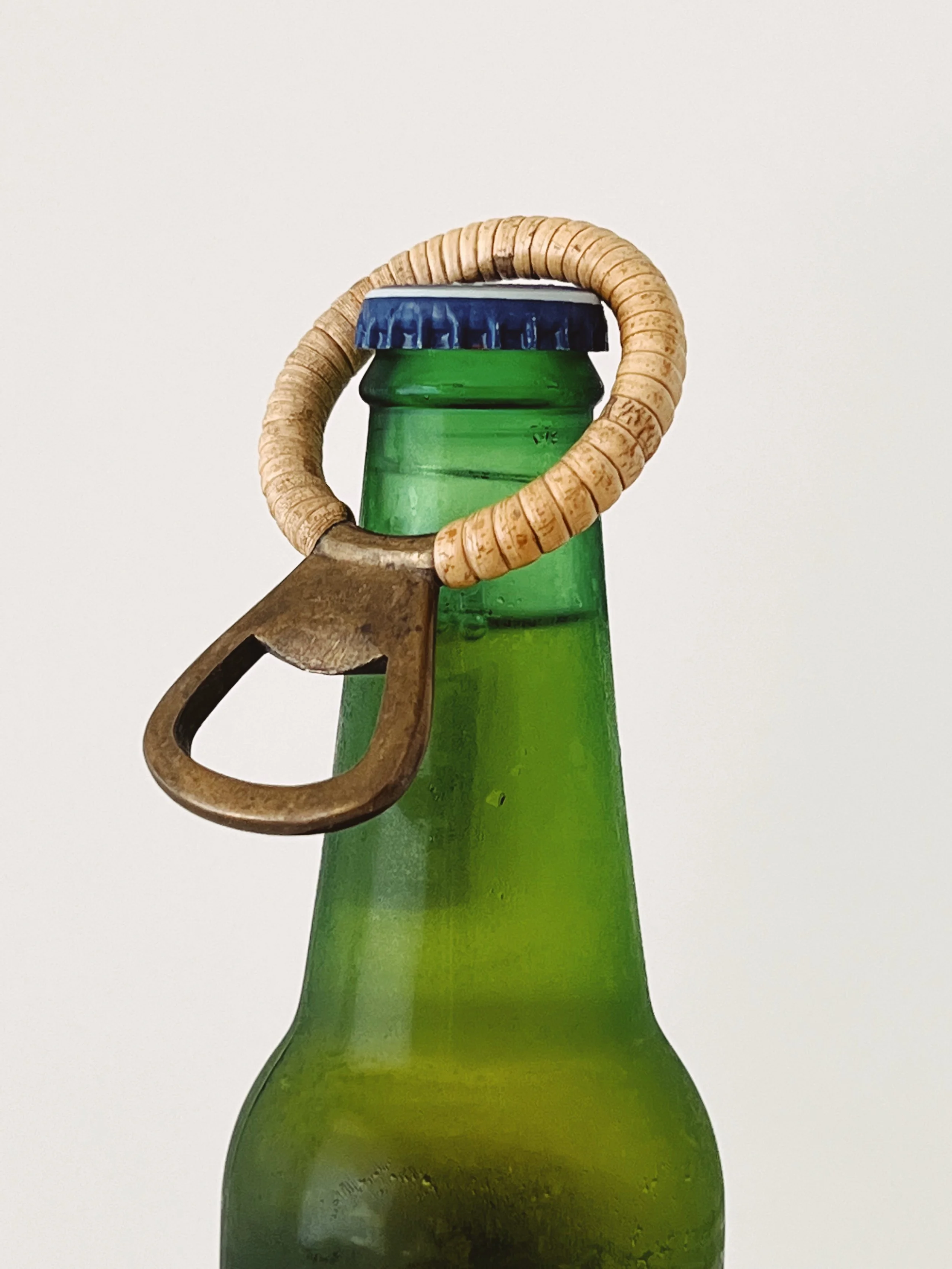 Close-up of a green glass bottle with a blue bottle cap, wrapped with a beige ring, and a rusty, old metal can opener hooked onto the ring, against a plain white background.