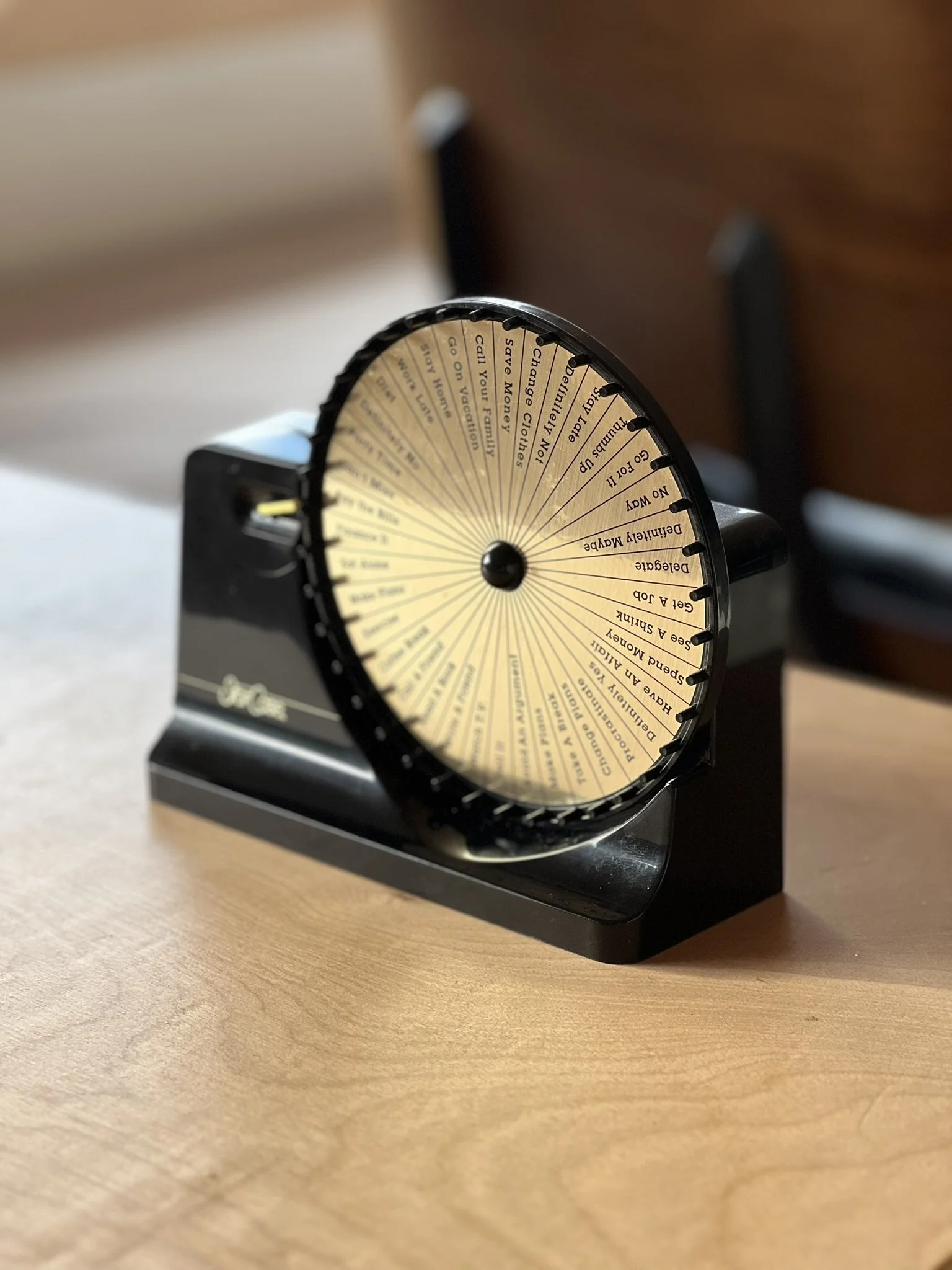 A vintage-style rotary dial wheel with instructions and phrases on a circular yellow background, mounted on a black stand, placed on a wooden surface.