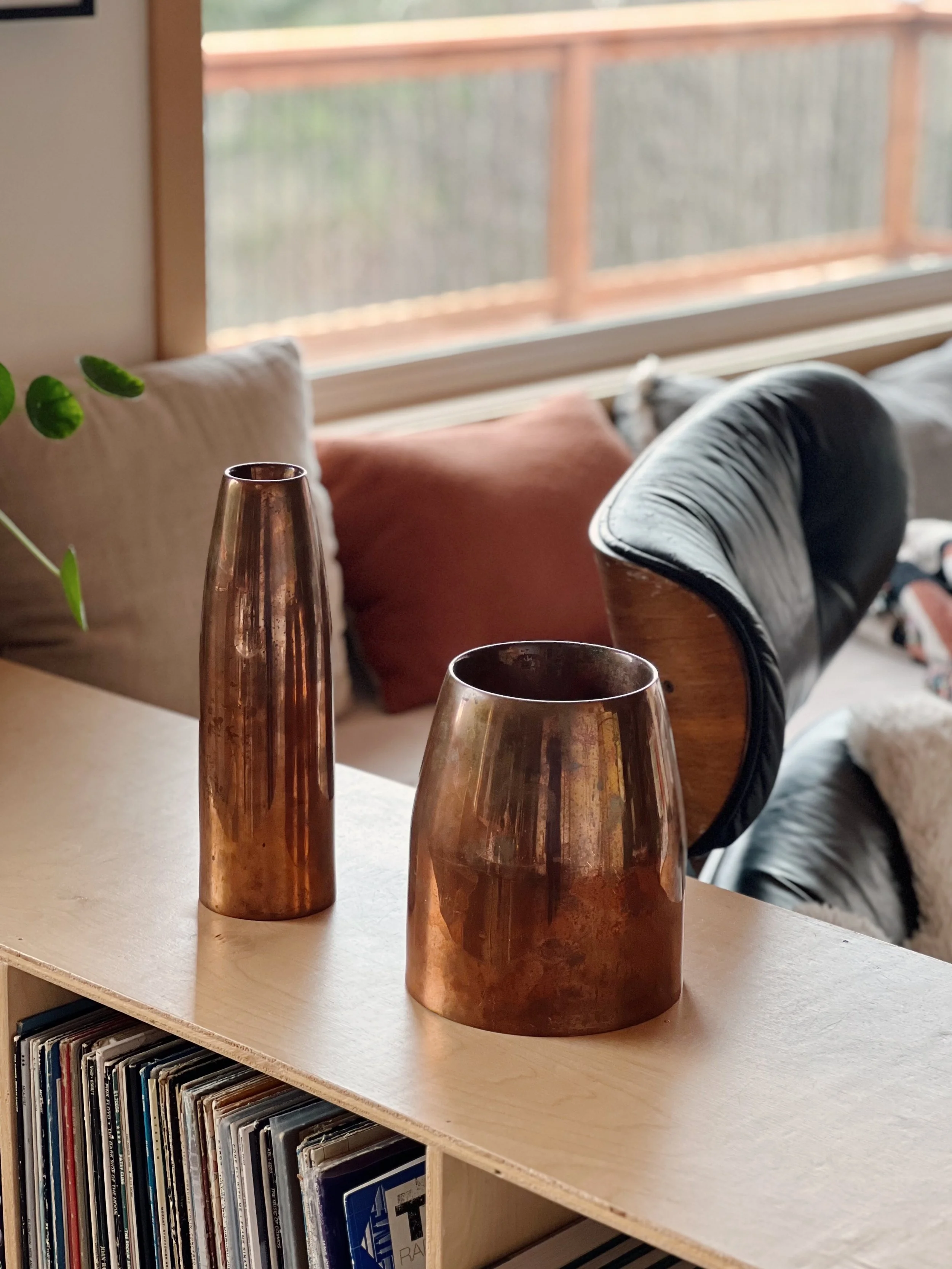 A close-up of three copper-colored vases of varying heights on a light-colored wooden shelf, with a collection of vinyl records underneath. In the background, there are pillow cushions and a window with a wooden railing outside.