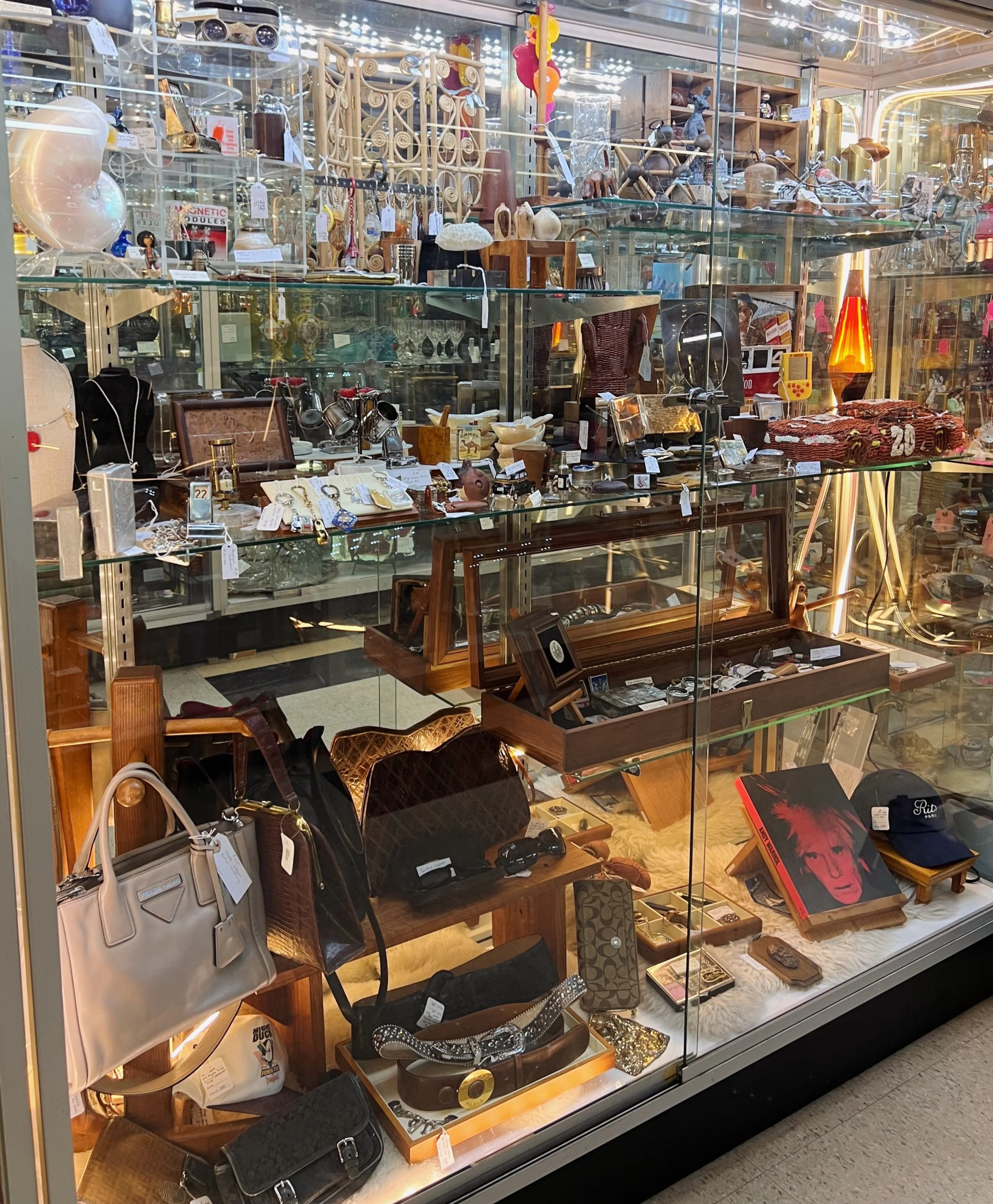 A glass display case filled with various vintage and collectible items, including handbags, jewelry, sunglasses, small sculptures, framed artwork, and decorative objects, arranged on multiple shelves in a store setting.