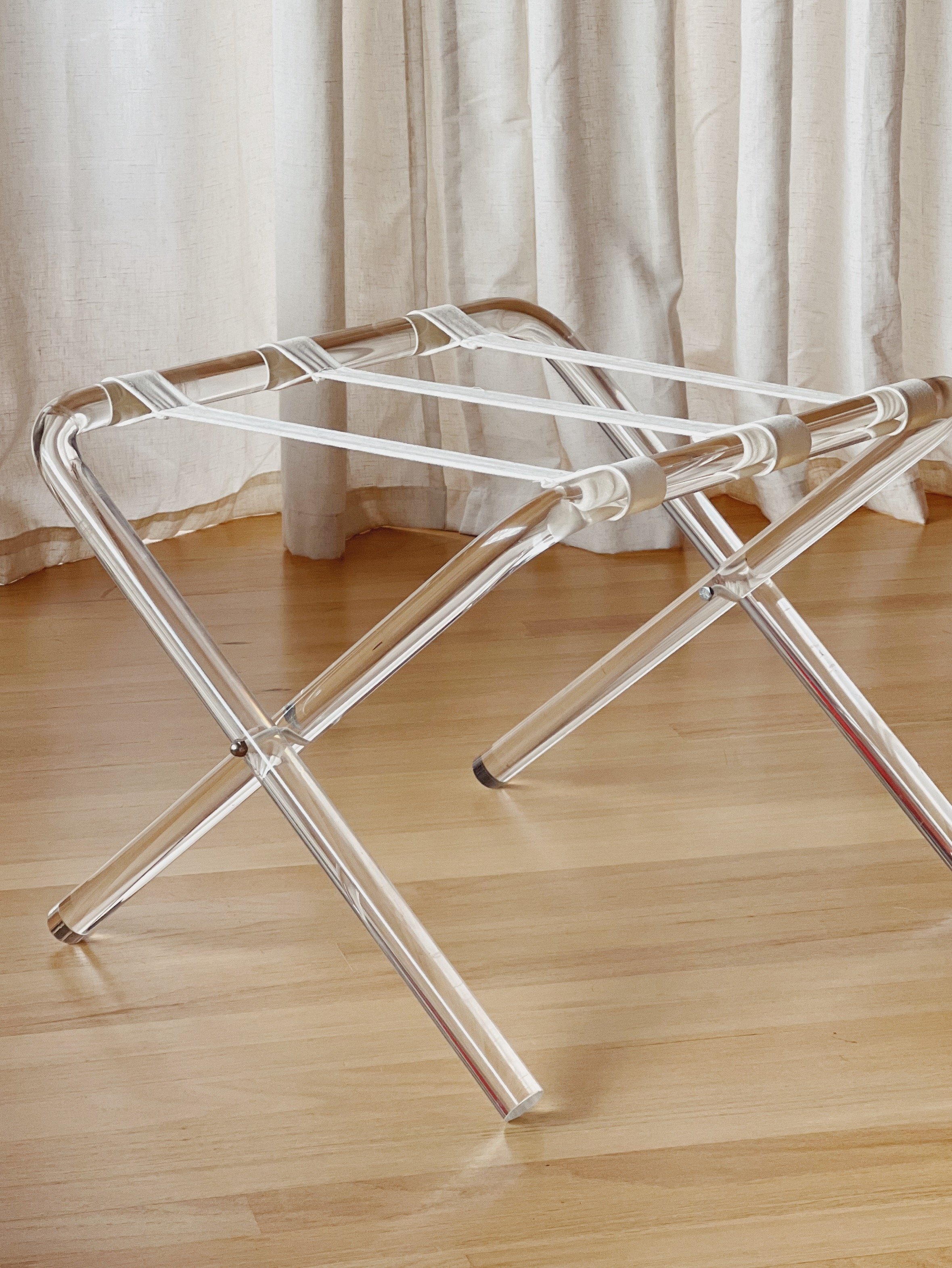 A foldable acrylic and metal chair placed on a wooden floor with beige curtains in the background.