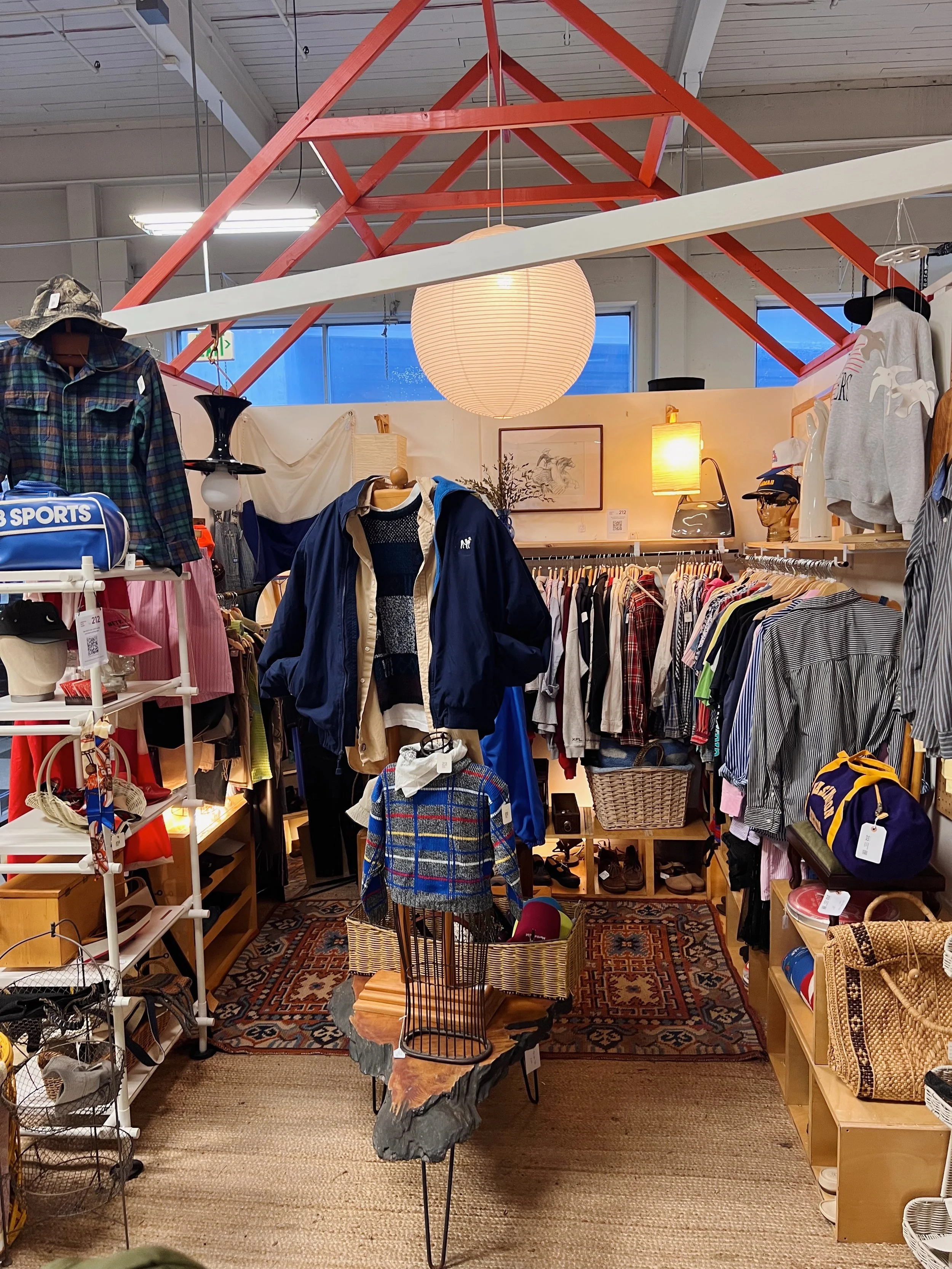 Interior of a thrift store with clothing, accessories, and home decor items displayed on shelves and racks, a large paper lantern hanging from the ceiling, and a patterned rug on the floor.