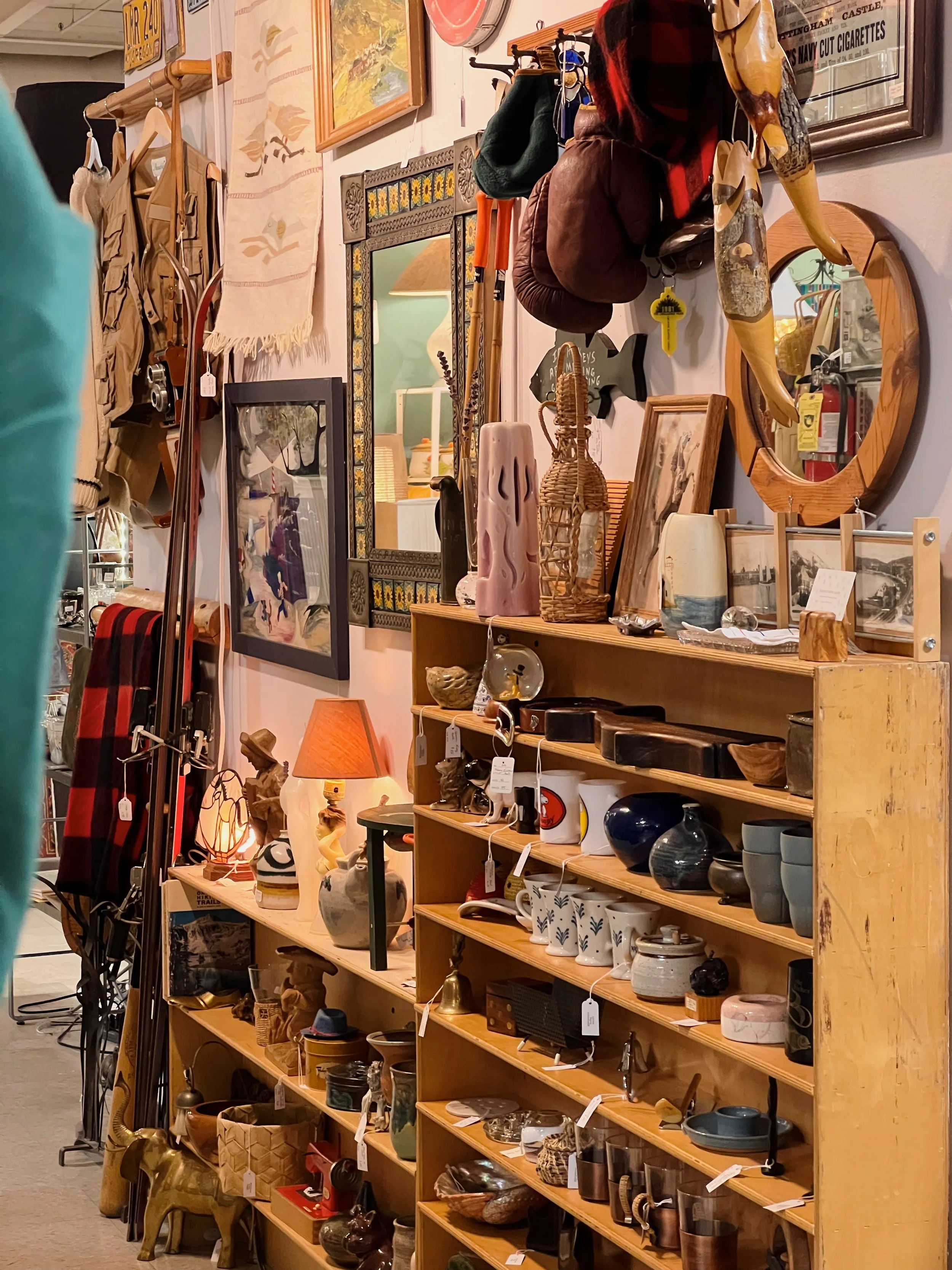 A display shelf filled with various home decor items including vases, bowls, and sculptures. Behind the shelf, there are mirrors, framed pictures, and hanging clothing and accessories. The background shows part of a store interior with other decor items and a glimpse of the store's furnishings.