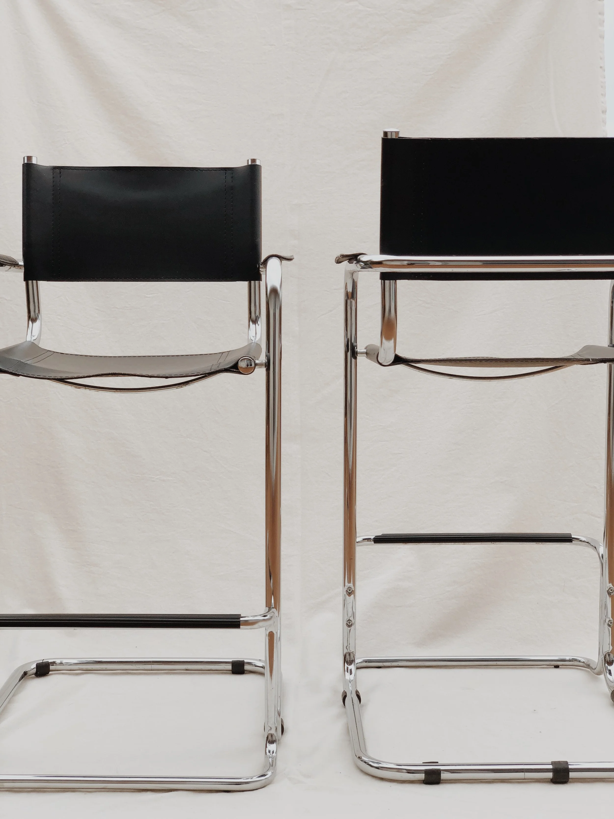 Two modern chairs with black leather backs and chrome frames, set against a white wall background.