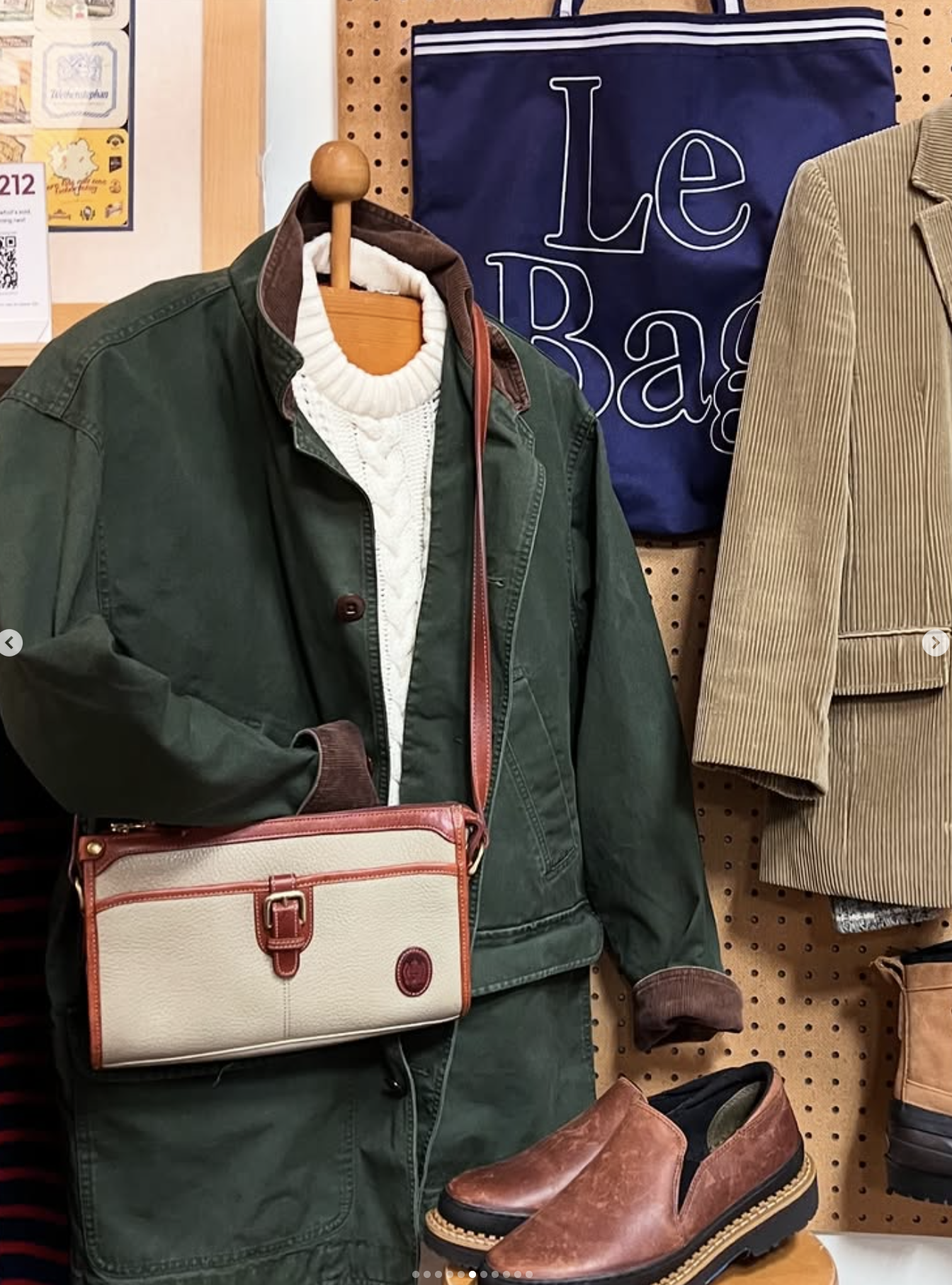 Display of men's clothing including a green jacket over a white sweater on a mannequin, a beige corduroy blazer, and brown leather shoes, with a blue sign that reads 'Le Bar' in the background.