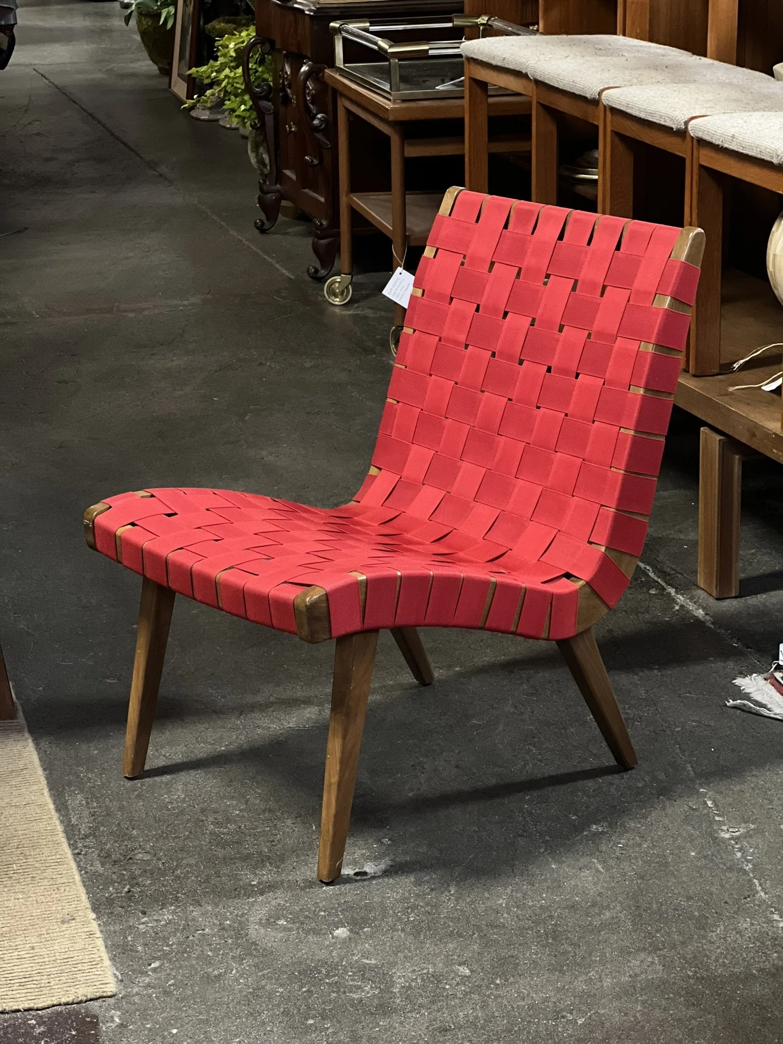 A red woven-style chair with wooden legs is positioned on a dark flooring surface, with a beige area rug to its left. Behind the chair, there are wooden tables and shelves displaying various furniture items, including a small wooden cart with wheels 