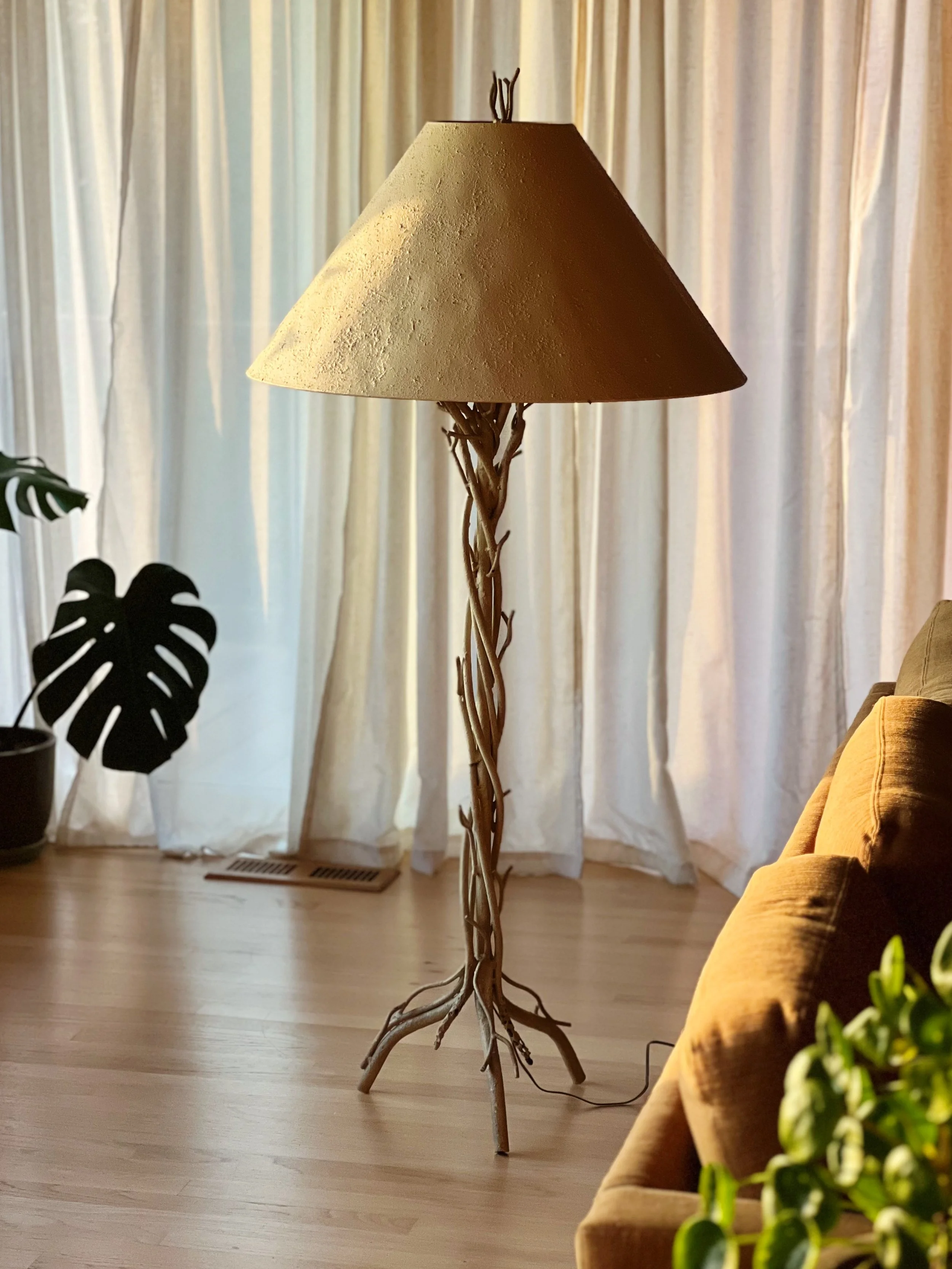 A floor lamp with a beige lampshade and a base made of intertwined branches, standing on a hardwood floor in front of cream-colored curtains. To the left, there is a potted plant with large, dark green leaves, and on the right, part of a mustard-colo