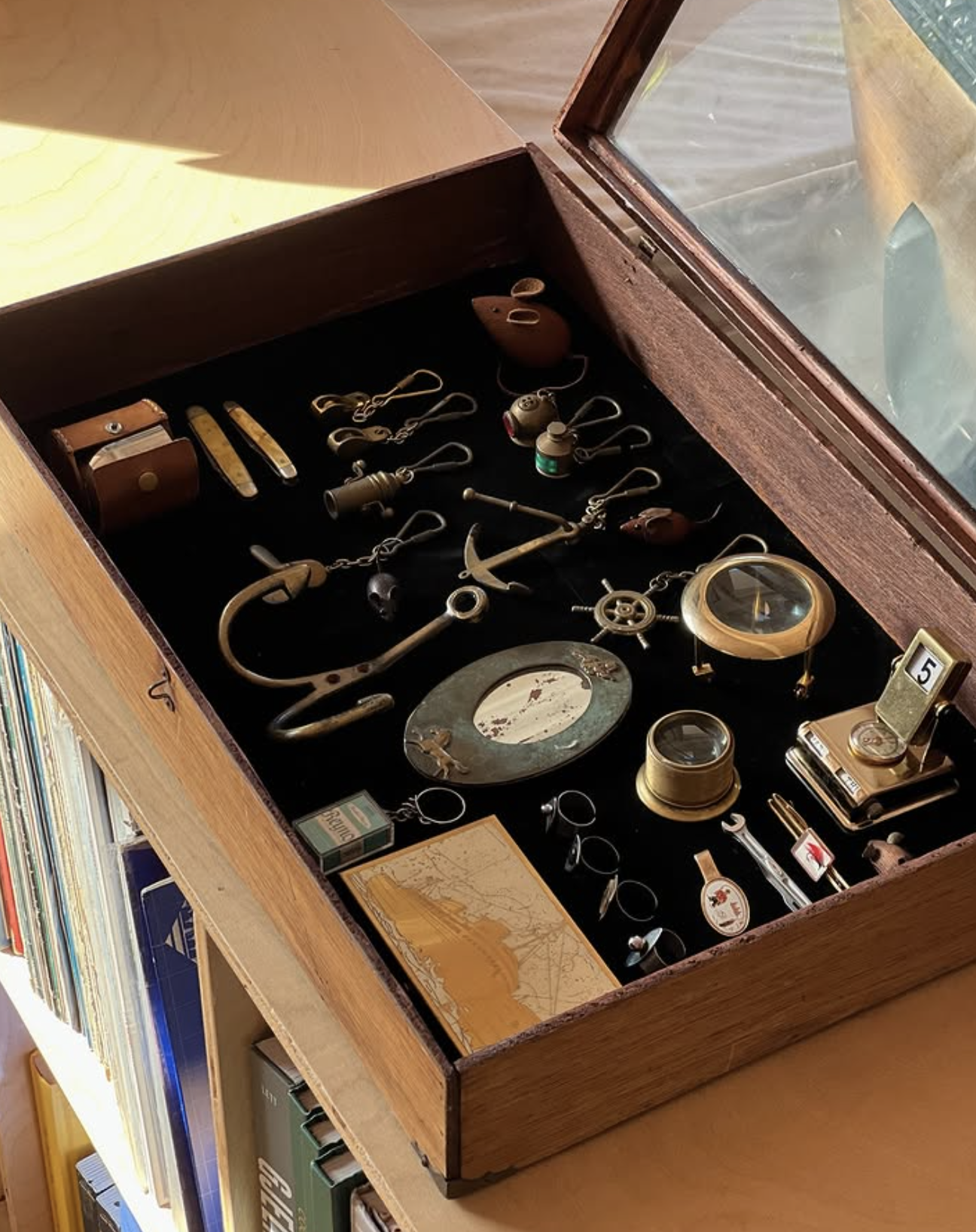 A wooden display box containing vintage jewelry and small collectibles, including rings, watches, and decorative items, with a glass lid and located on a wooden table beside books.