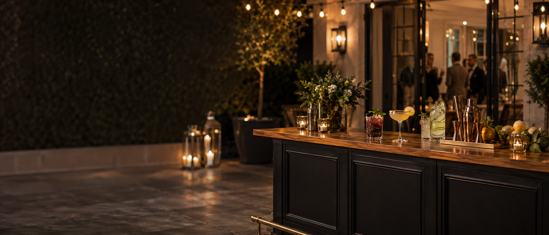An outdoor evening social gathering with a wooden bar, various cocktails, floral arrangements, and candlelight. People are seen in a warmly lit interior through open glass doors.