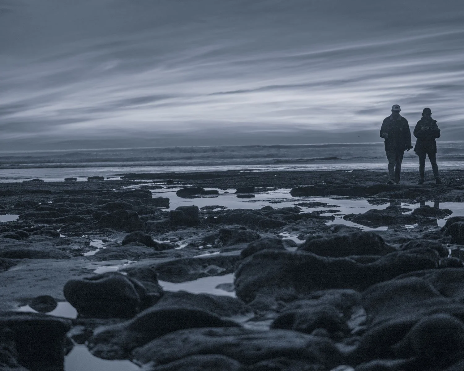 Two people walking along a rocky beach at dusk or dawn with an overcast sky and waves in the background.