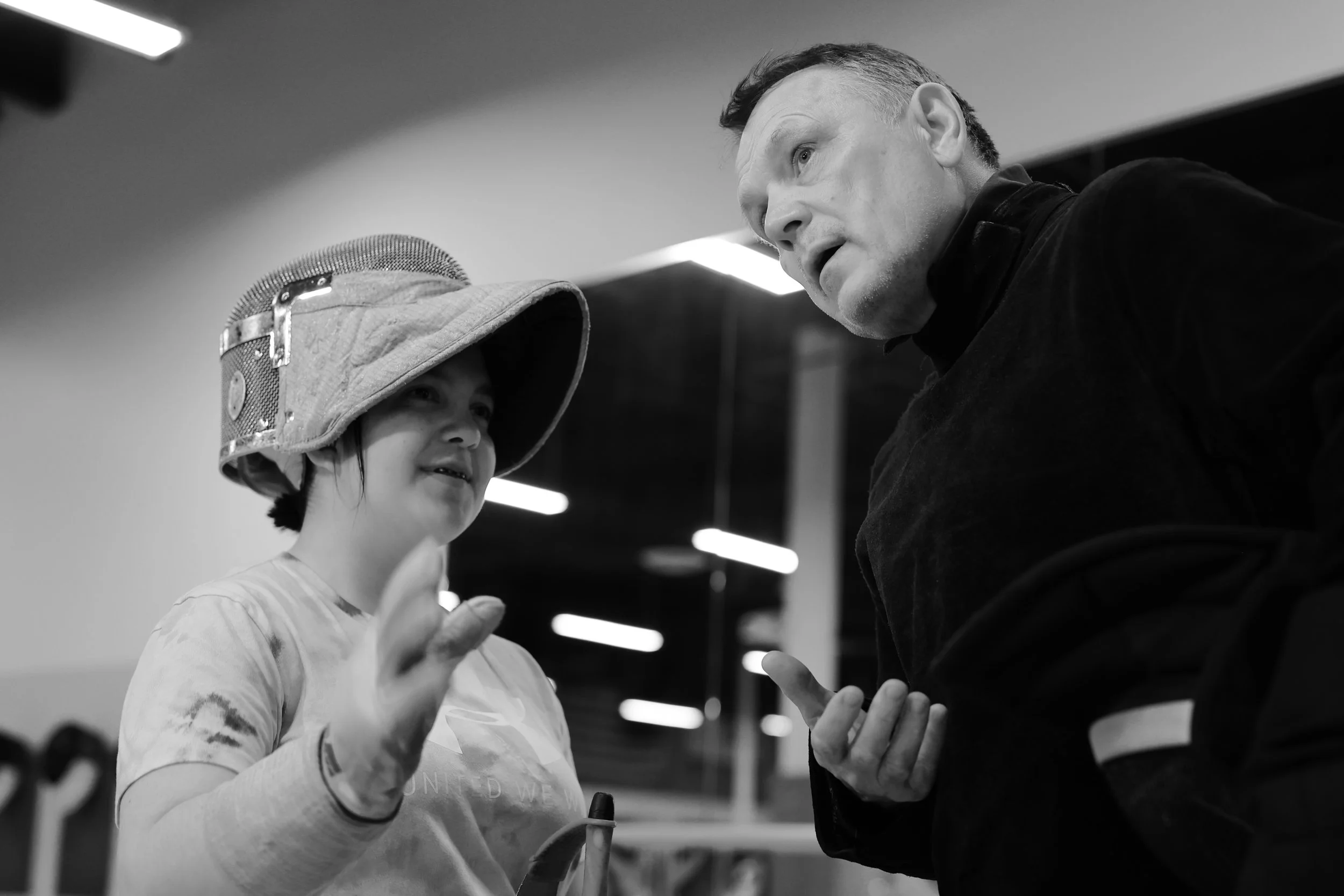 A woman wearing a hat and a patterned shirt talking to a man with short hair and a dark top. The man is listening intently and they appear to be engaged in a conversation. The background is blurred indoors with bright lights.