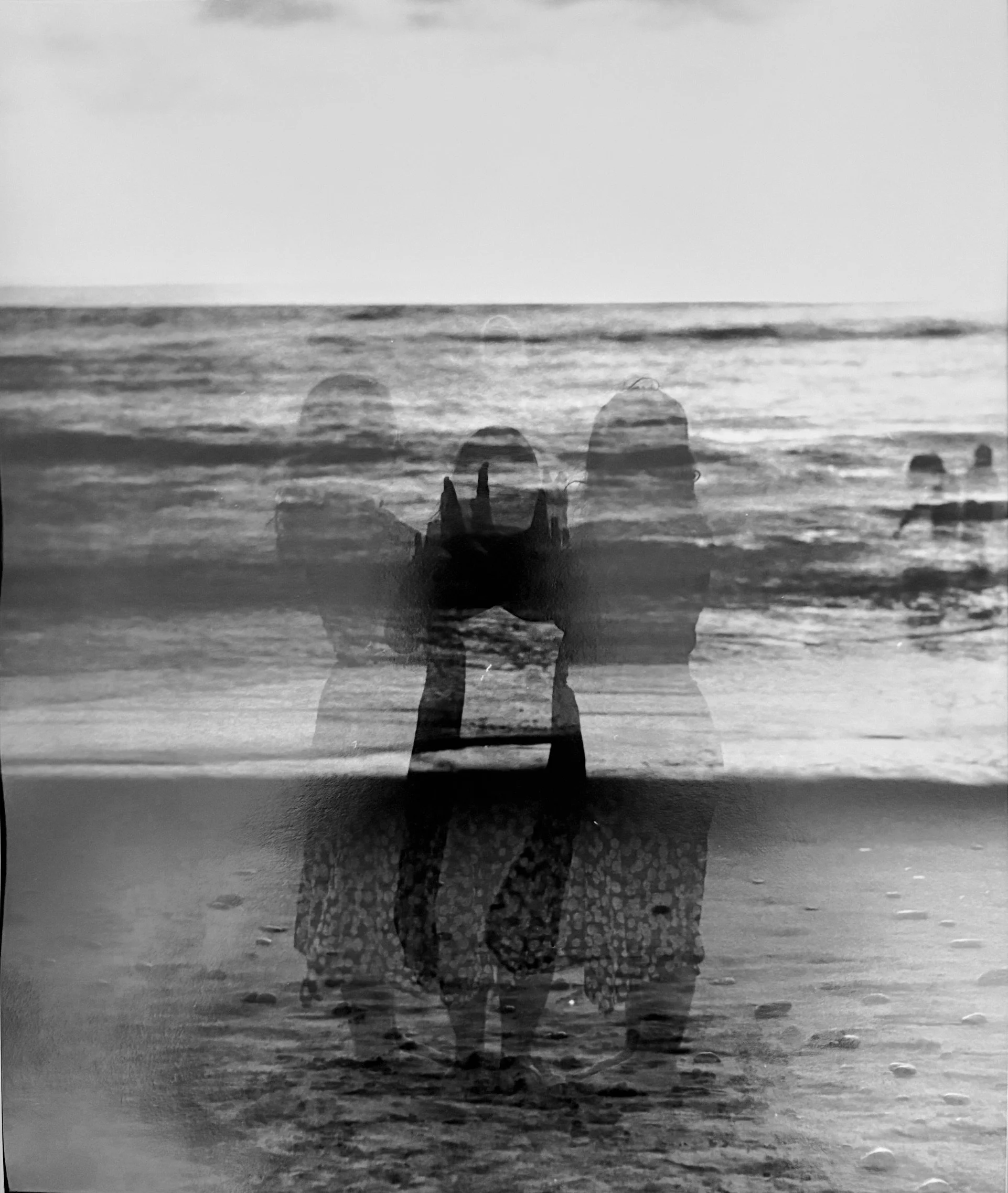 Black and white photo of two people standing in water on a beach, with ocean waves and sky in the background, slightly transparent, creating a layered double exposure effect.
