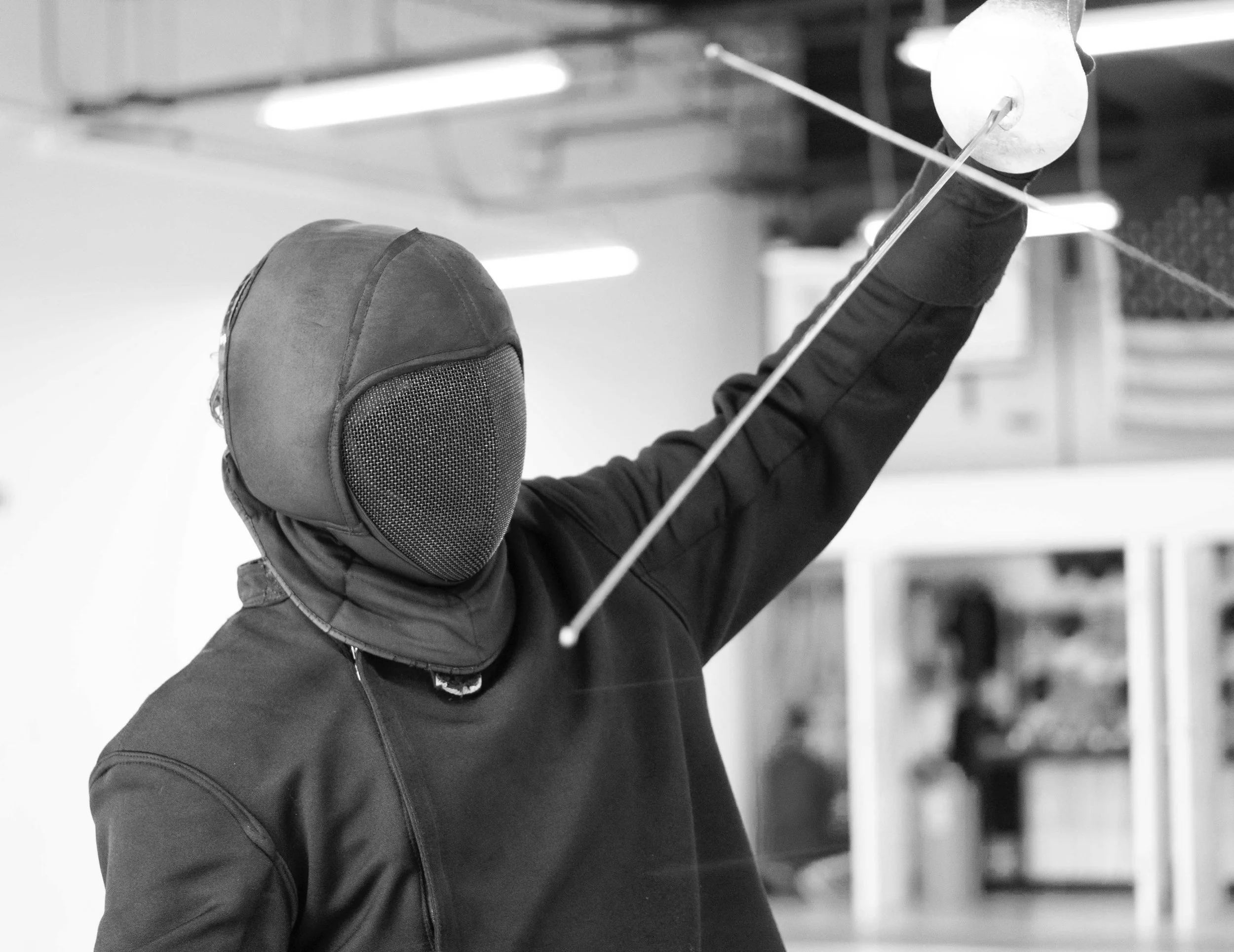 A person fencing in an indoor training facility, wearing a protective mask, black jacket, and gloves, holding a fencing foil.