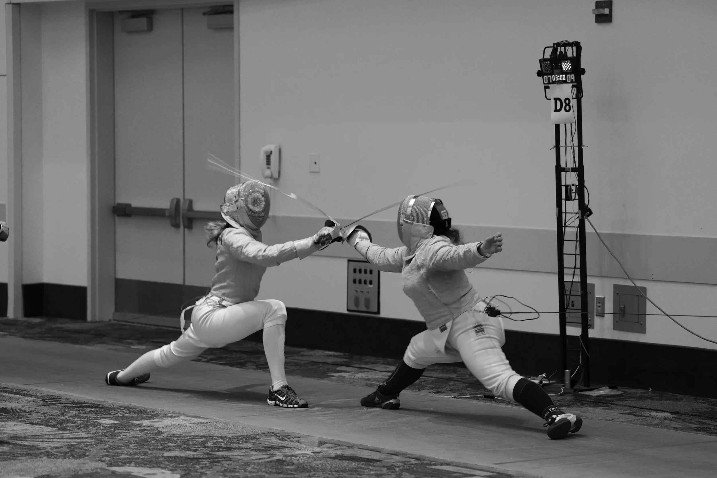 Two women fencing indoors, wearing fencing gear and masks, engaged in a foil or epee match, with a digital scoreboard in the background.