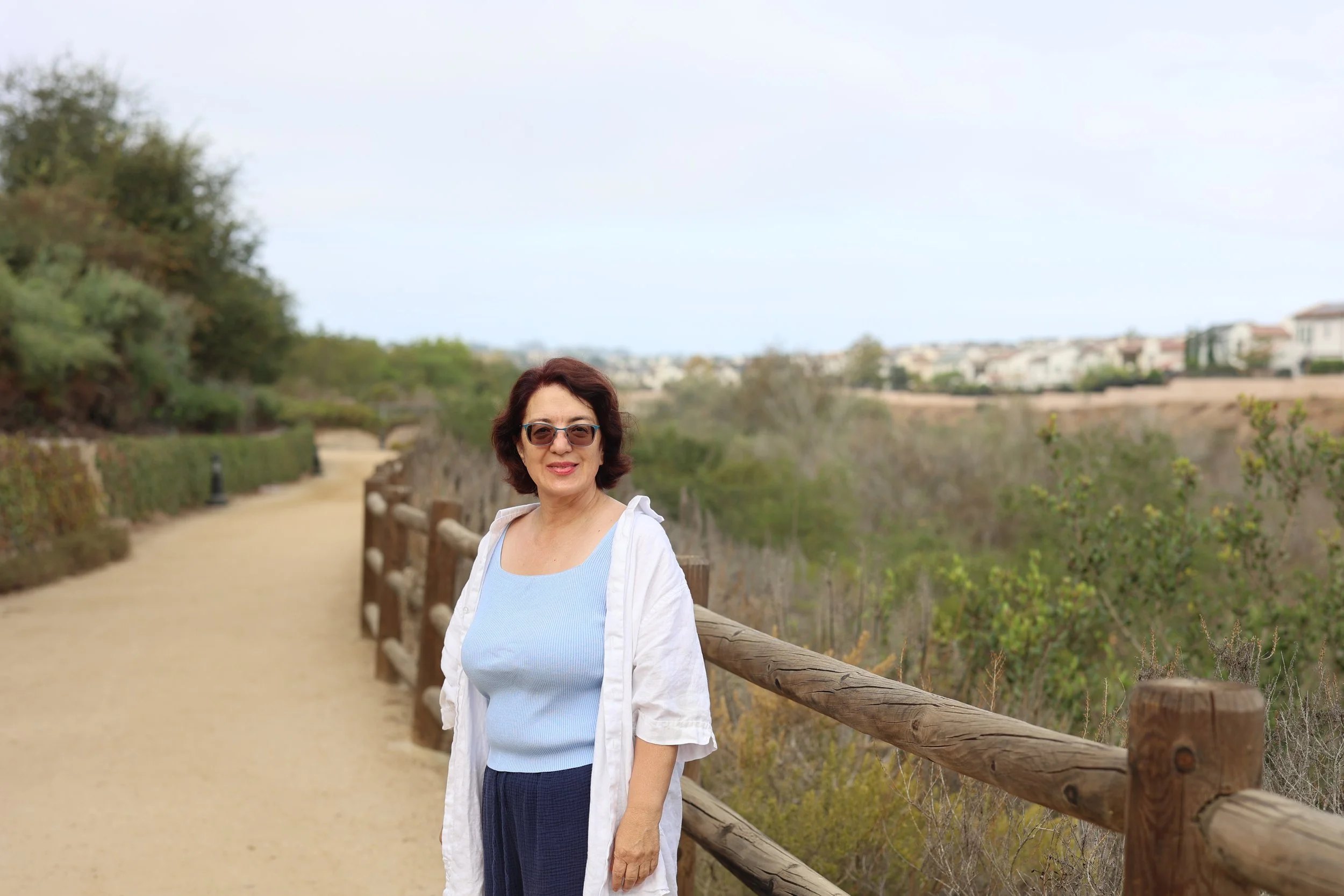 A woman with short dark hair and sunglasses standing outdoors next to a wooden fence along a nature trail with greenery and distant houses in the background.