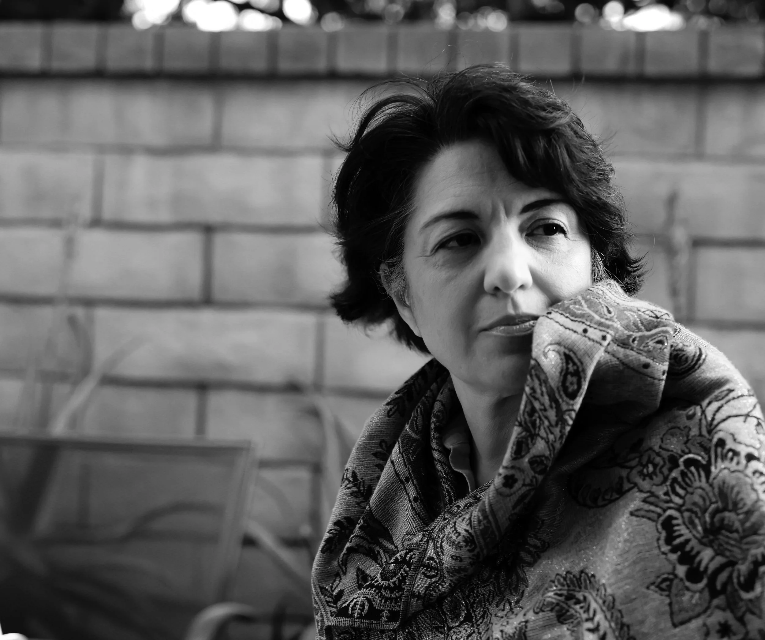 Close-up of a woman's face with short curly hair, wearing a patterned scarf and a jacket, leaning against a brick wall.