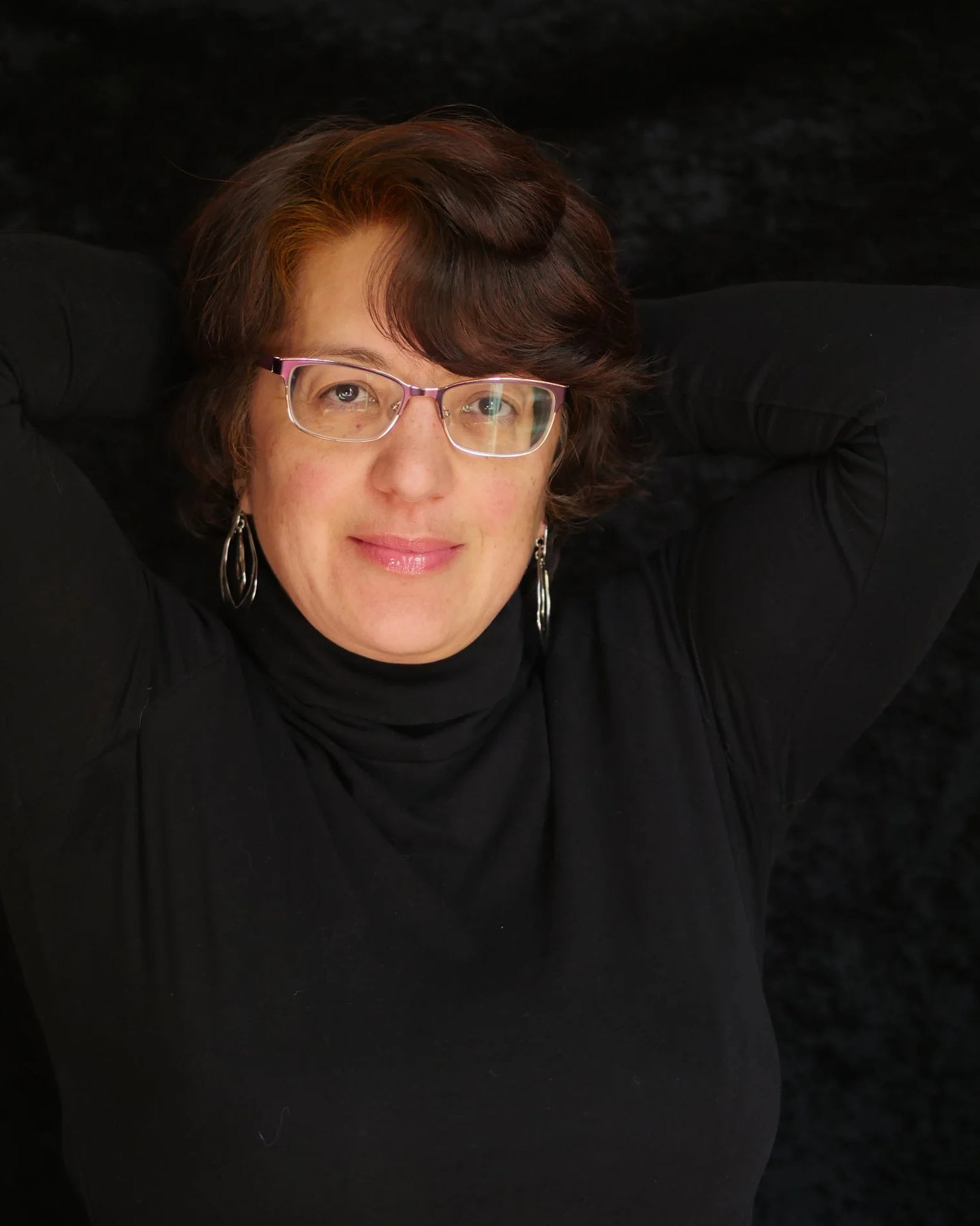 A woman with short, reddish-brown hair wearing glasses and silver earrings, resting her arms behind her head, smiling at the camera against a dark background.