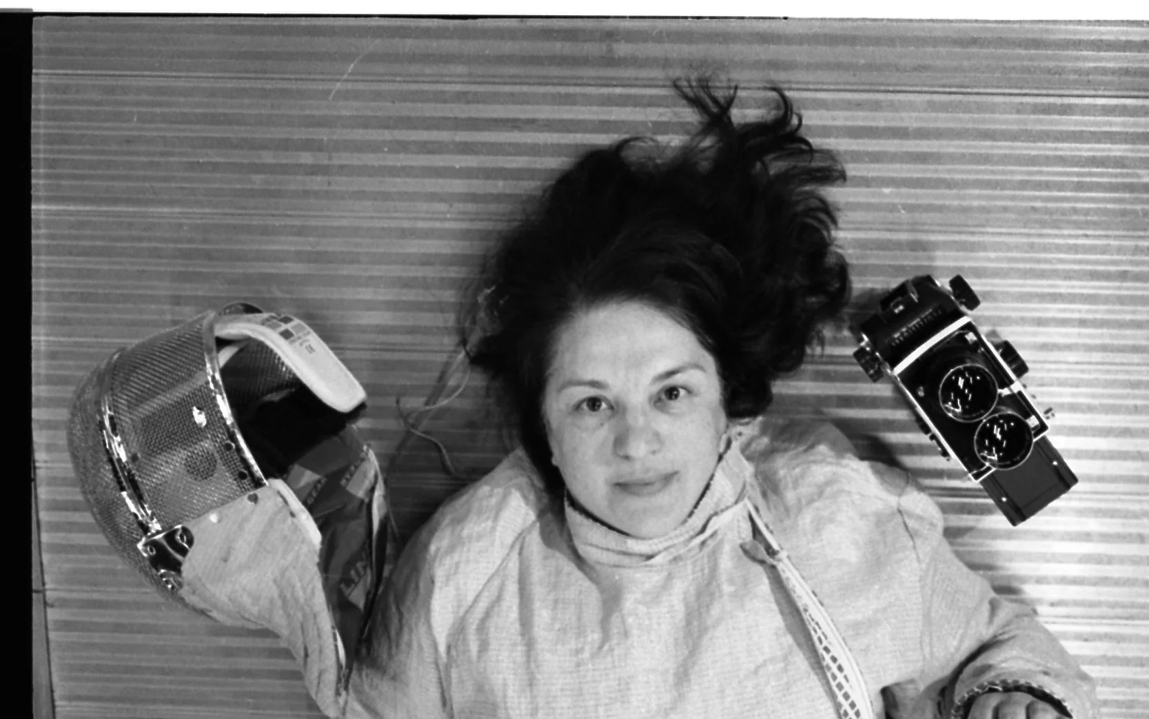 A woman with long, curly hair lying on a striped surface, looking up at the camera. She is wearing a light-colored jacket and has a relaxed expression. Beside her on the surface are a helmet and a vintage camera.