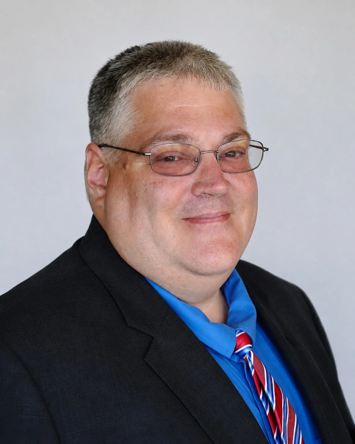 A man with short, gray hair and glasses wearing a black suit, a blue dress shirt, and a red, white, and blue striped tie, smiling against a plain background.