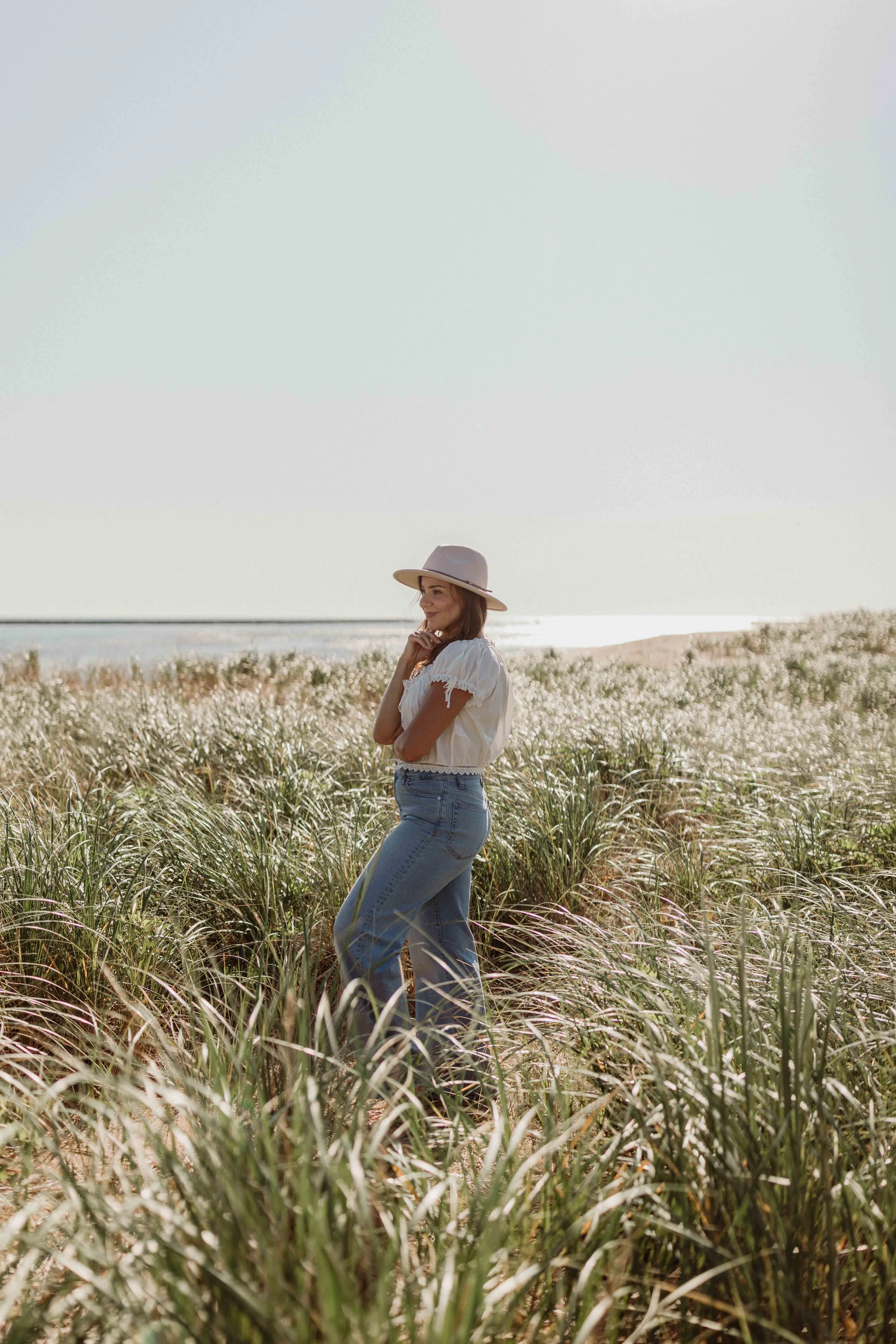 Woman in a white blouse and jeans standing in a field of tall grass near the coast, wearing a wide-brimmed hat, smiling, with the ocean in the background.