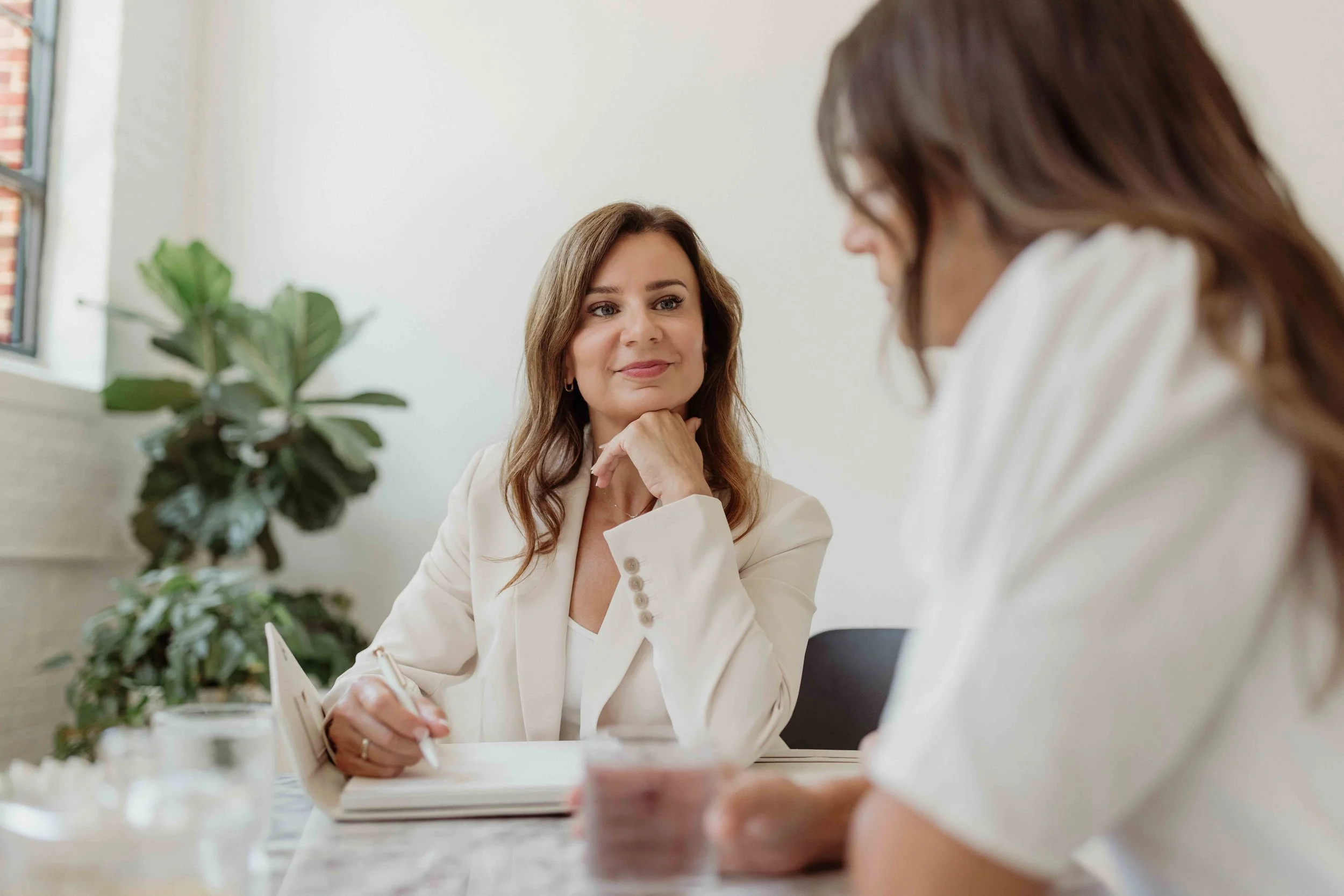 A woman in a white blazer sitting at a table, listening to and engaging with another woman who is out of focus in the foreground, in a bright room with a plant and a window.