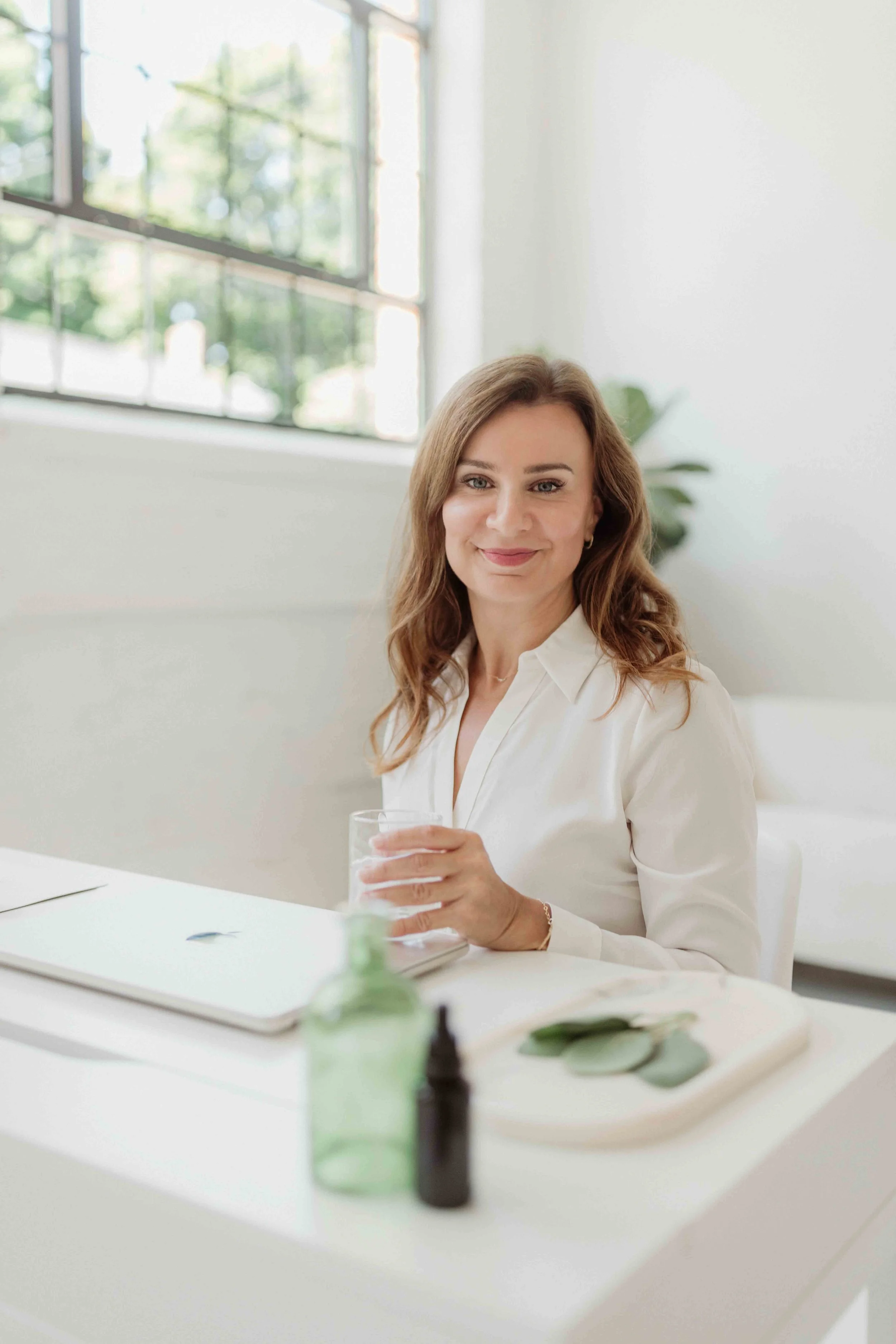 A woman with light skin and brown hair, smiling, sitting at a white table in a bright room with large windows and greenery outside, holding a glass of water.