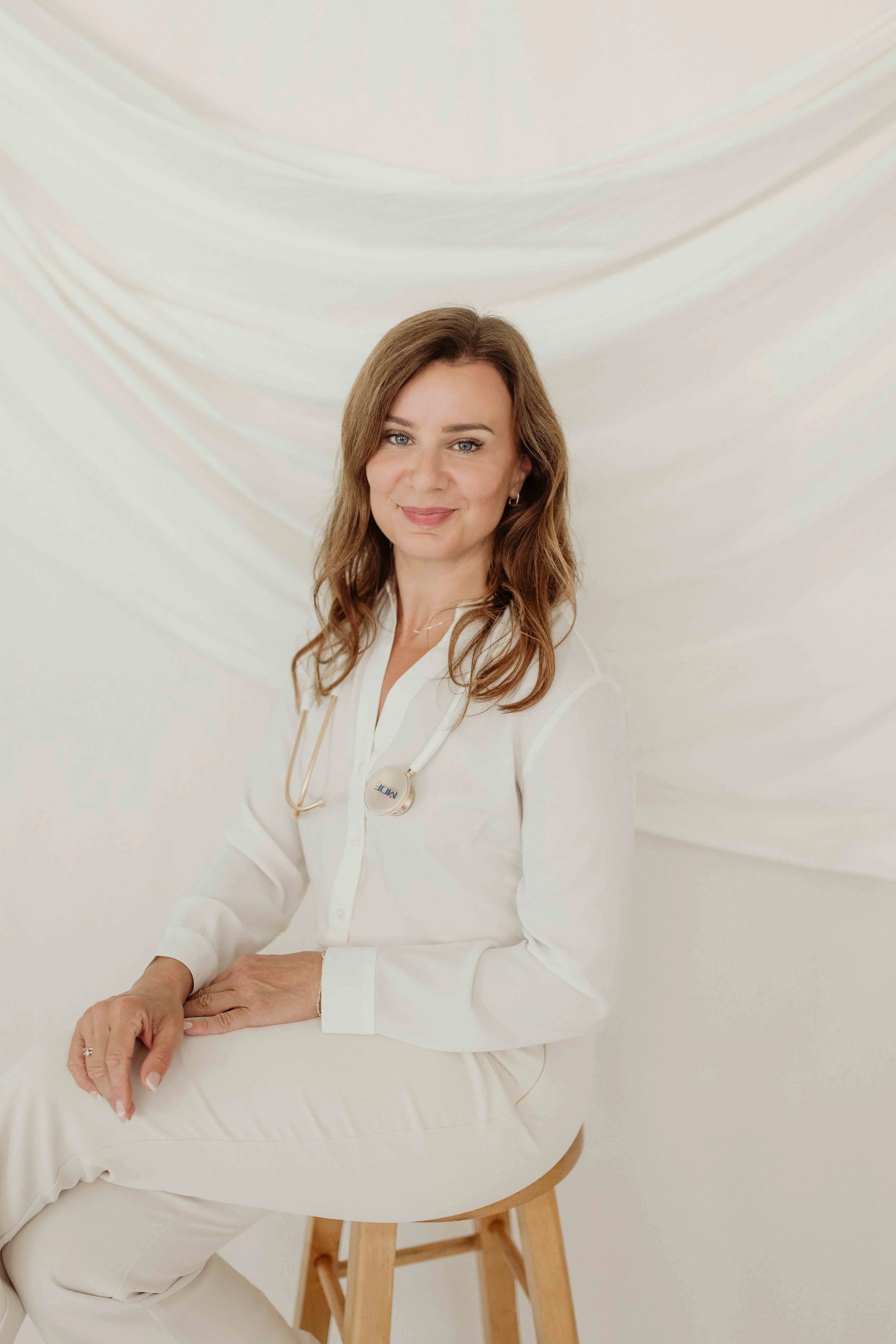 A woman with long, wavy brown hair and blue eyes, wearing a white medical coat and a stethoscope, sitting on a wooden stool. The background is a white draped fabric.