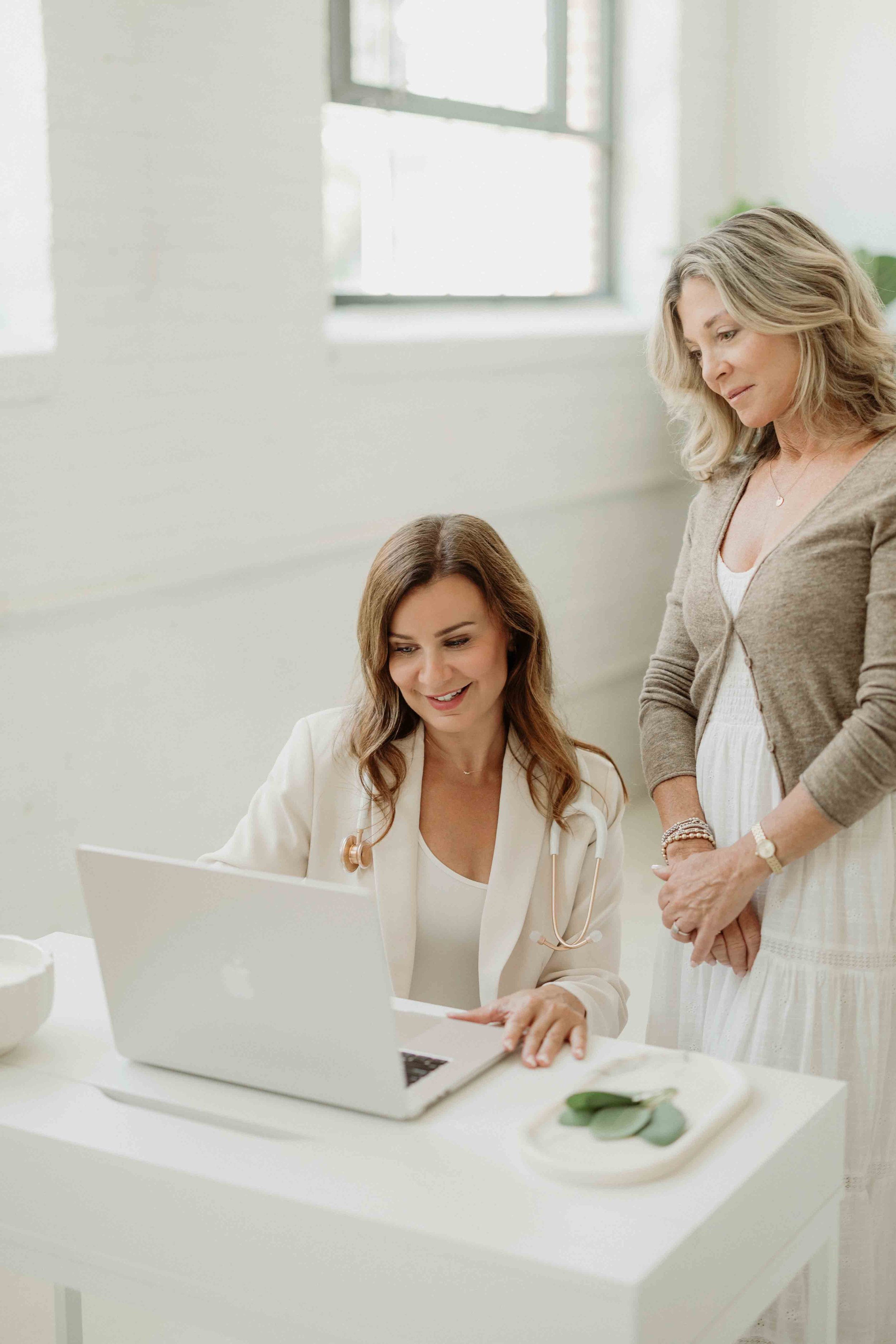 A female doctor with a stethoscope on her neck is sitting at a white desk with a laptop, looking at the screen. An older woman, possibly a patient, stands beside her, looking at the laptop screen with a slight smile in a bright, modern medical office with large windows.