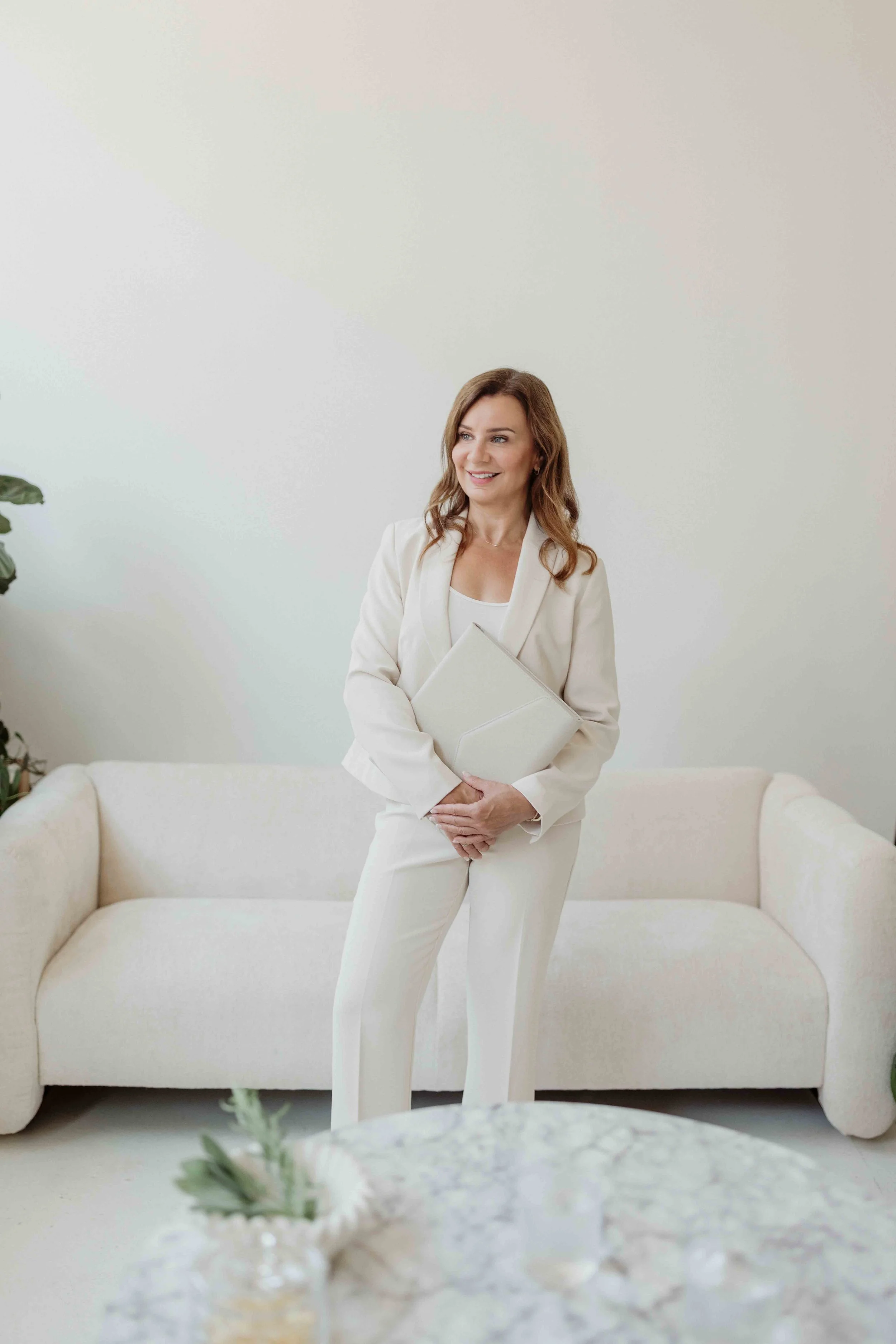A woman dressed in a white suit, standing in a bright, minimalistic room, holding a folder and smiling. There is a white sofa behind her and a small plant on a table in the foreground.
