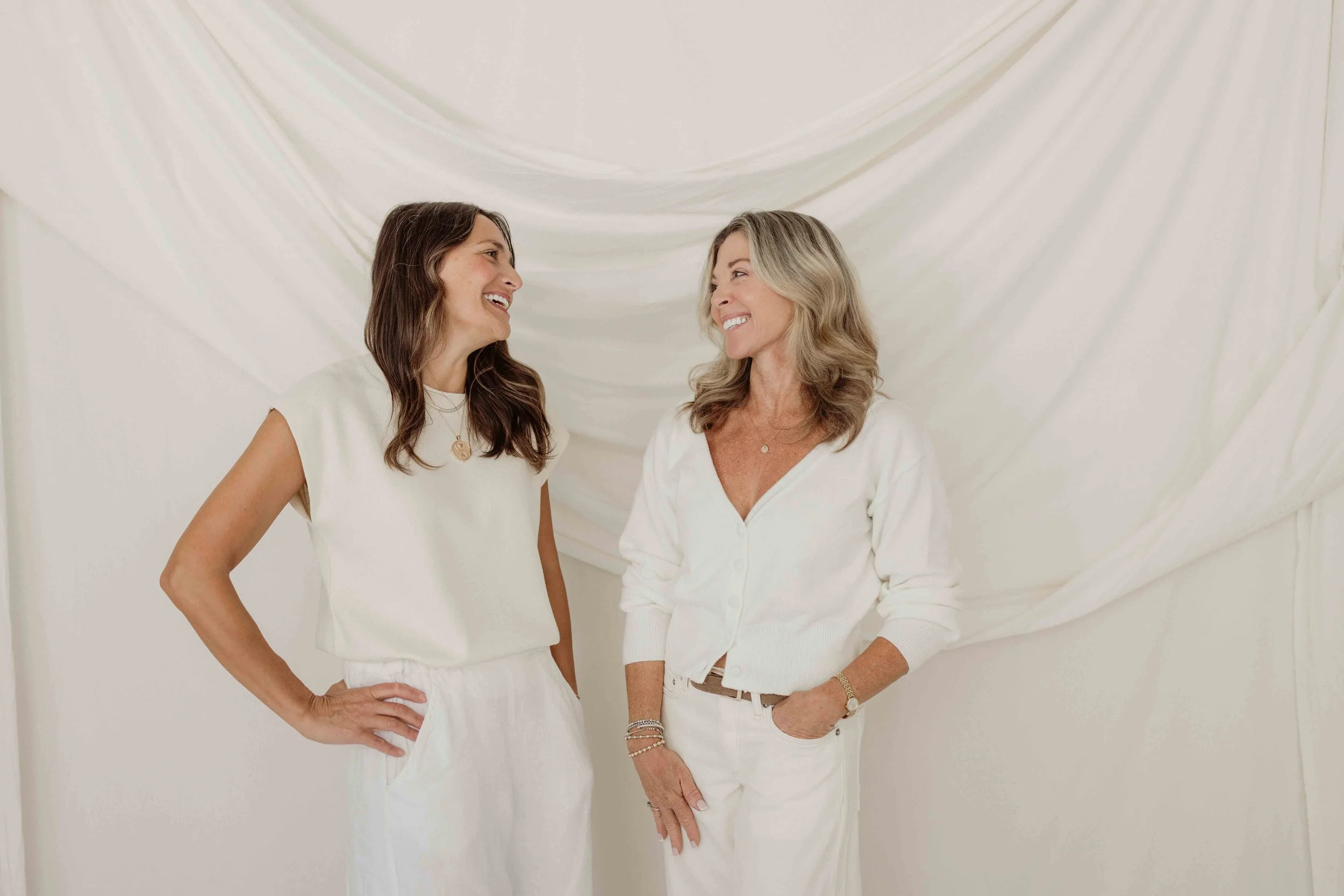Two women standing and smiling at each other in front of a white draped fabric backdrop, dressed in white casual clothes.