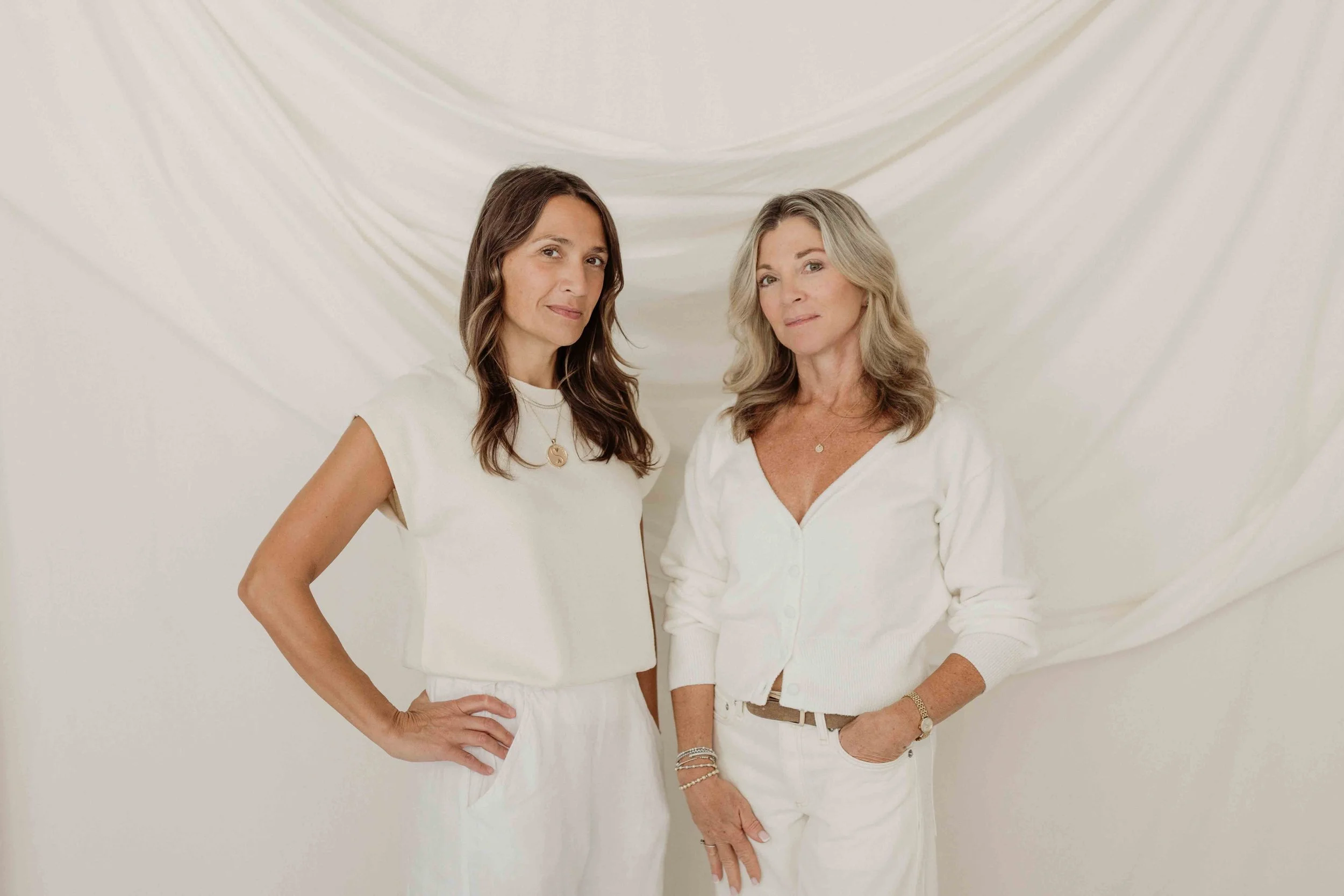 Two women standing side by side against a white fabric background, dressed in white clothing, looking at the camera.