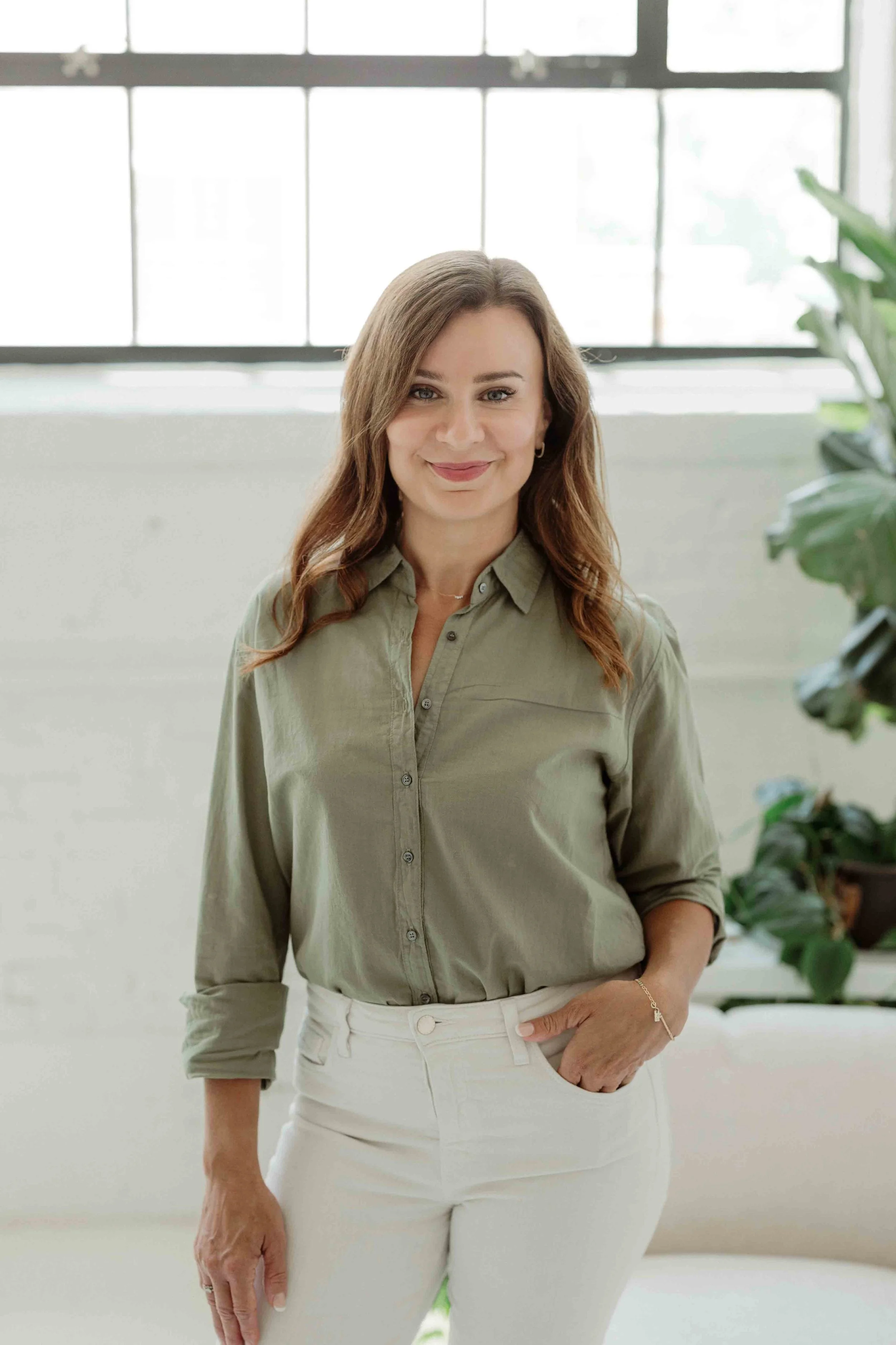 A woman with brown hair, wearing a light green button-up shirt and white pants, standing indoors near a large window with sunlight and a plant visible in the background.