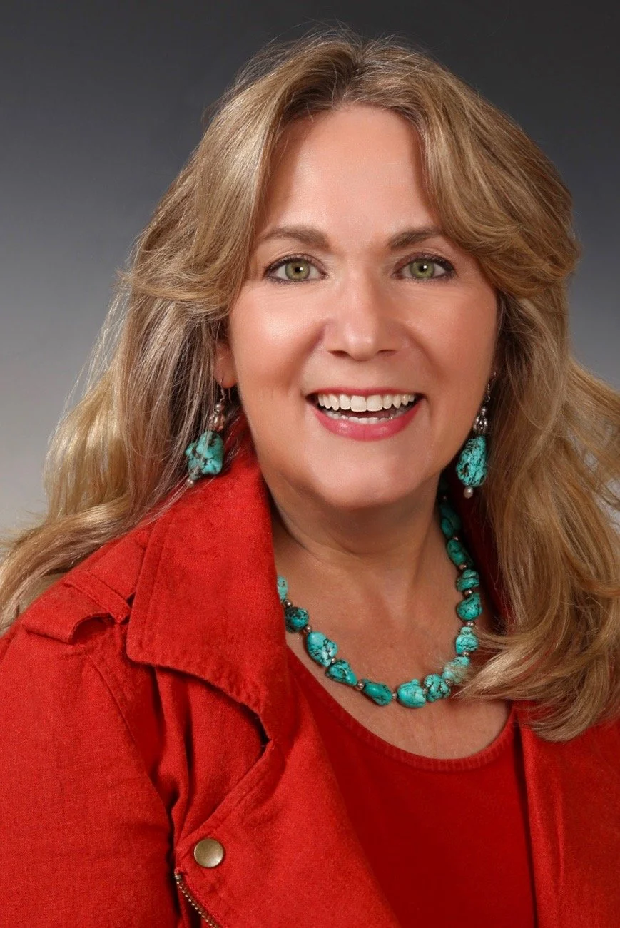 A woman with long blonde hair, green eyes, smiling, wearing turquoise jewelry and a red jacket against a dark gray background.