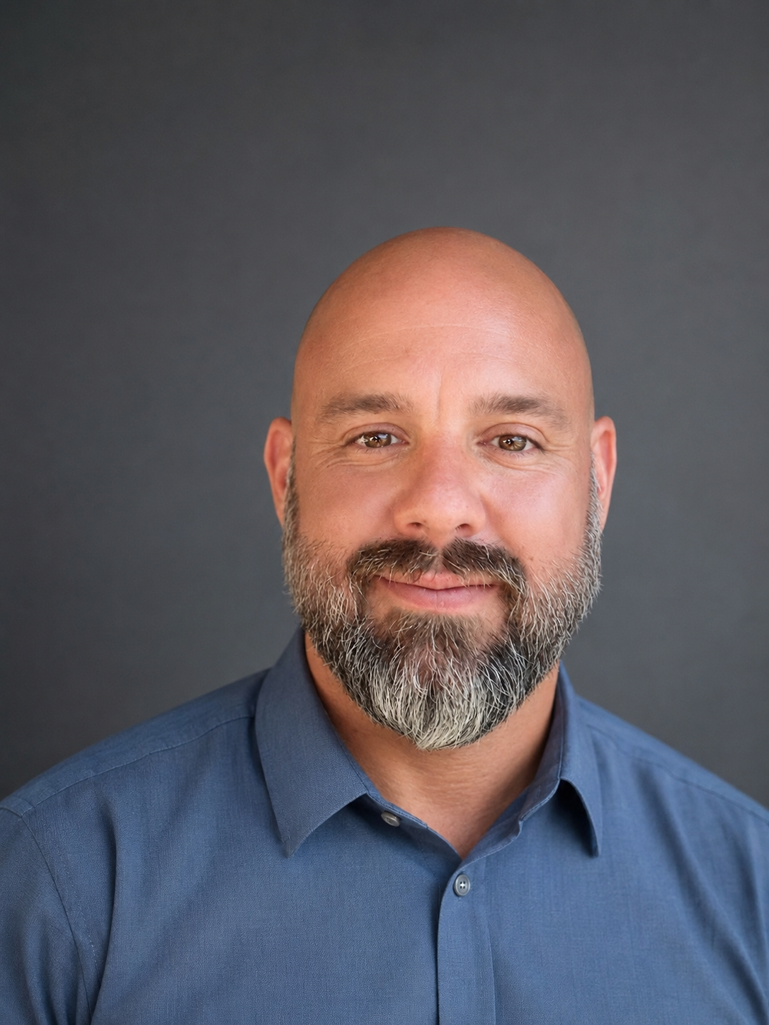 Headshot of a bald man with a beard, smiling, wearing a blue collared shirt, against a dark gray background.