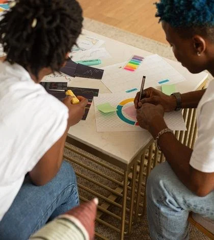 Two individuals working on a creative project at a table with papers, colored sticky notes, and markers