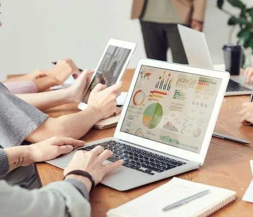 People working in a meeting room using electronic devices like a laptop and tablets with data charts and graphs on screen.