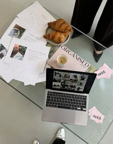 Desk with a laptop displaying a photo grid, papers, a bowl with croissants, and pink paper notes.