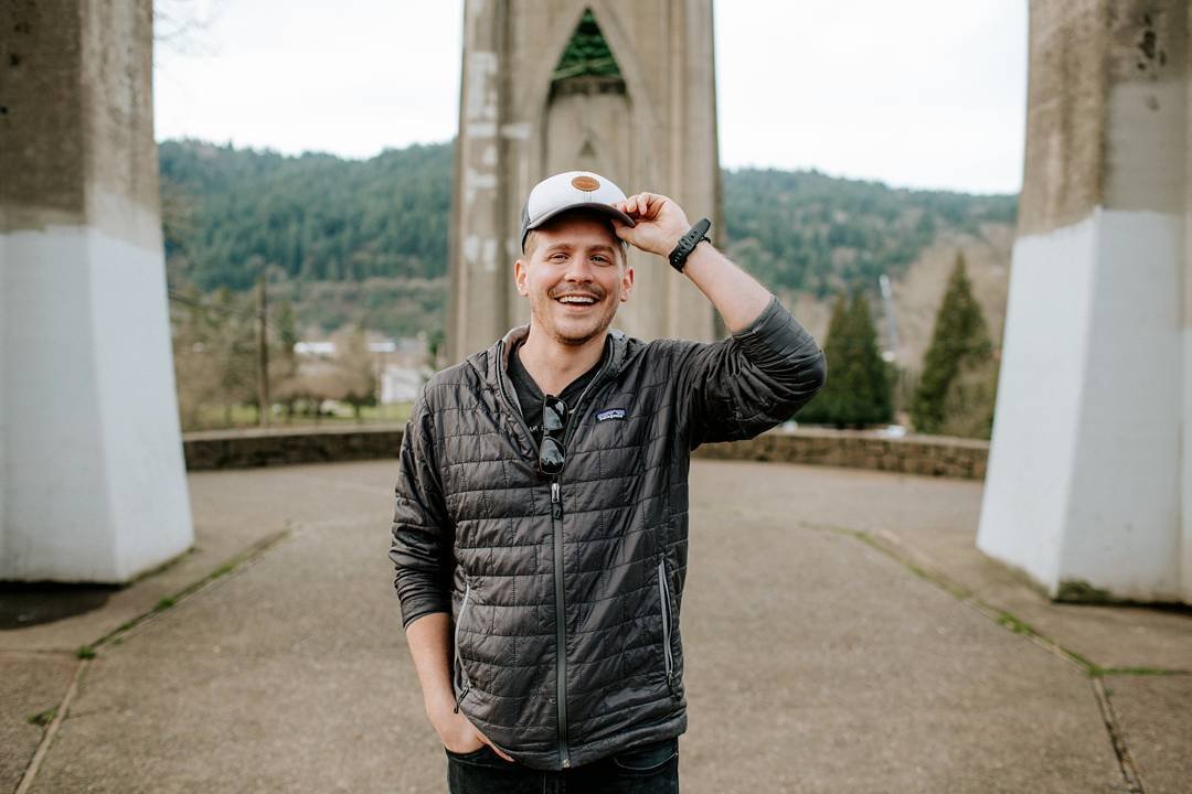 A young man with a beard and mustache smiling and wearing a black jacket, gray cap, and sunglasses hanging from his shirt, standing outdoors near a bridge with mountains and trees in the background.