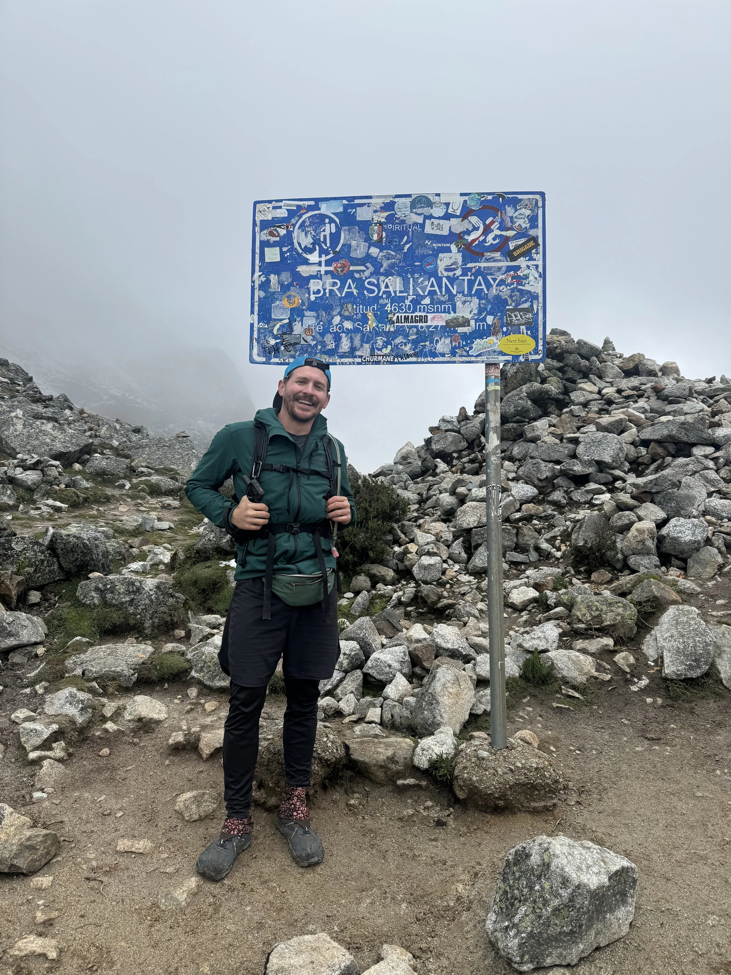 A man smiling and standing in a rocky mountainous area near a weathered sign covered with stickers and graffiti, indicating an elevation of 4630 meters above sea level.