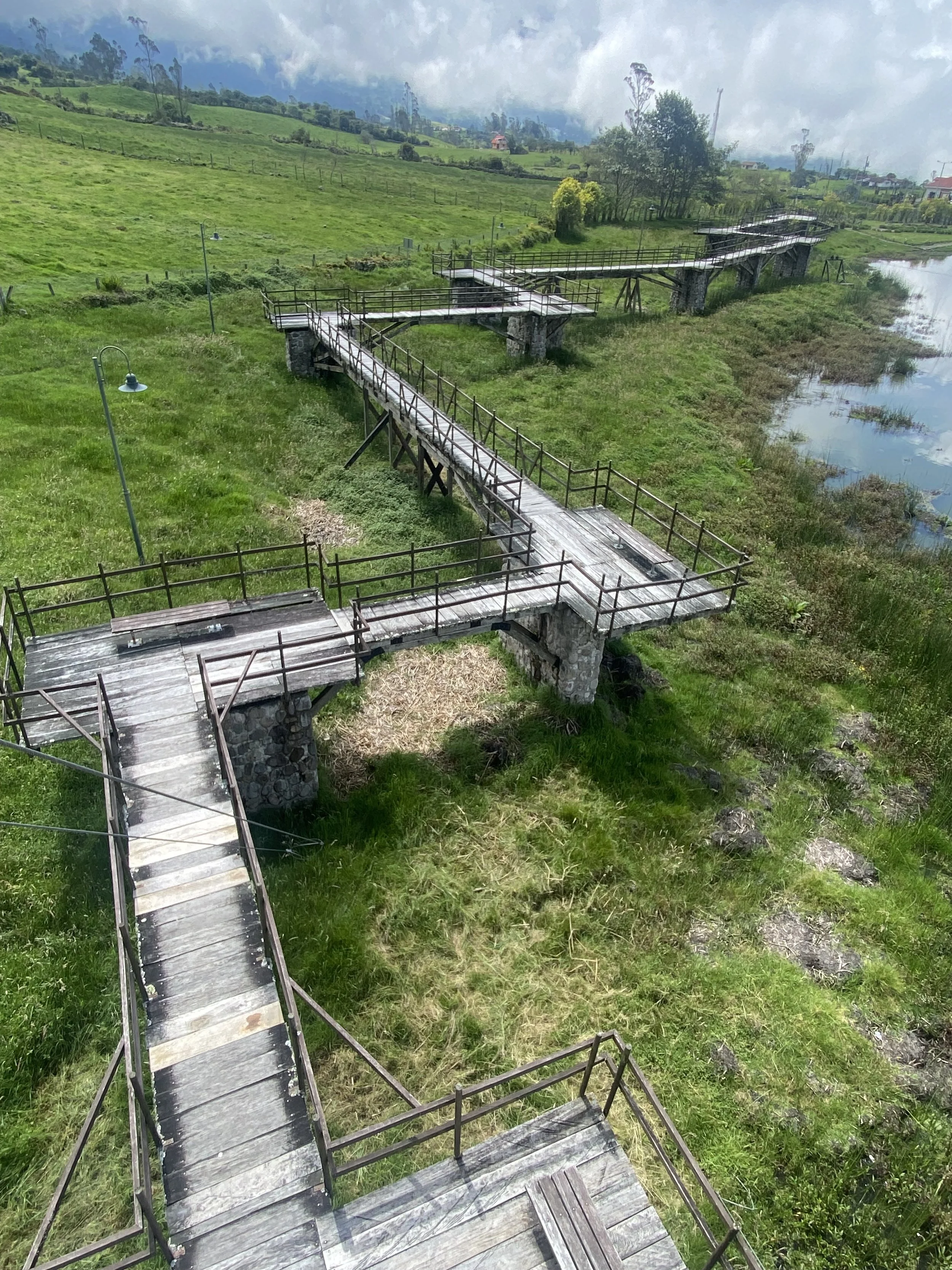 An elevated wooden walkway with metal railings winding over a grassy field and leading to a glass-bottomed bridge over a small pond in a lush green landscape with cloudy sky overhead.