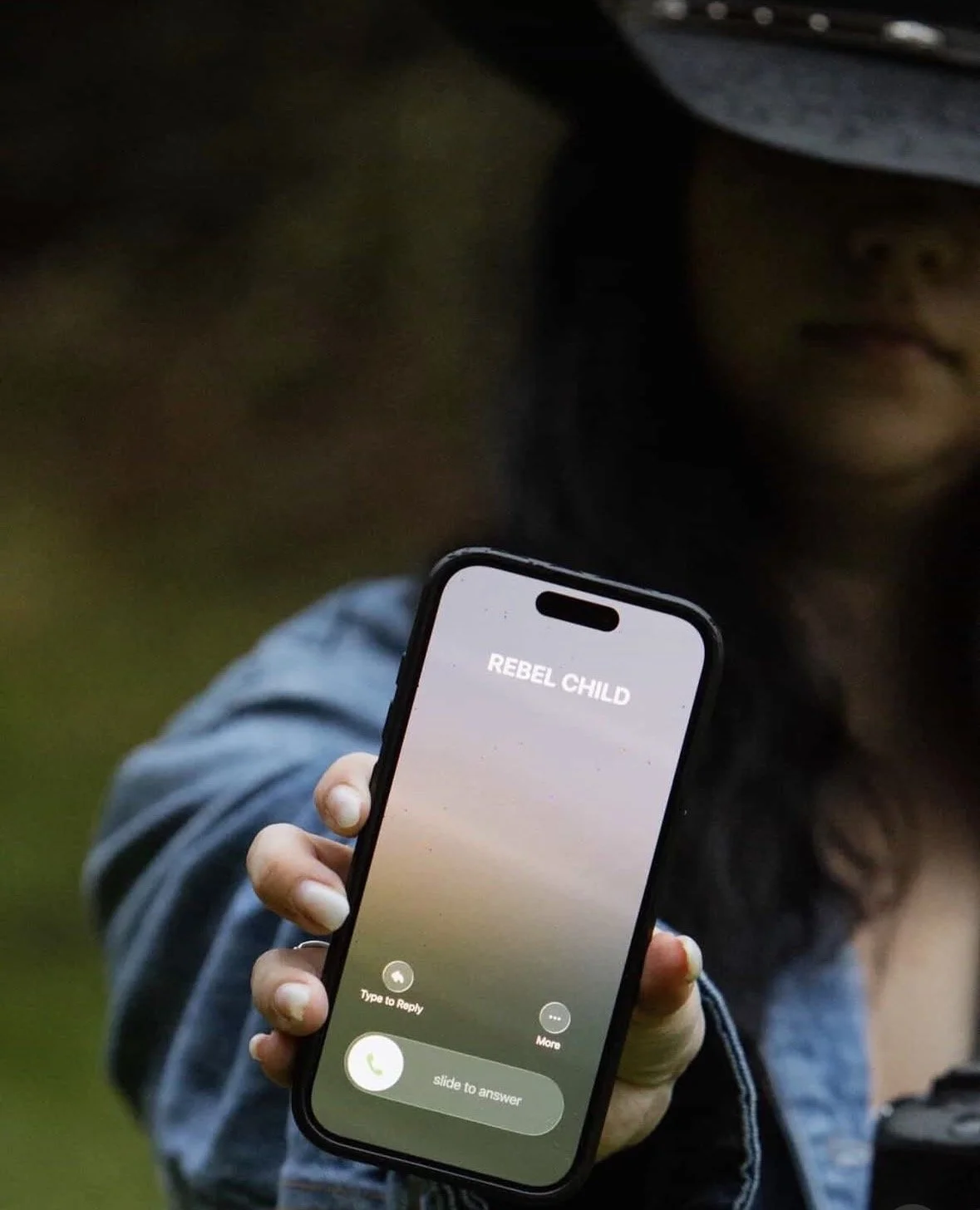 A person holding a smartphone displaying an incoming call from 'Rebel Child' outside during daylight. The person's face is partially visible, wearing a gray hat and clothing.