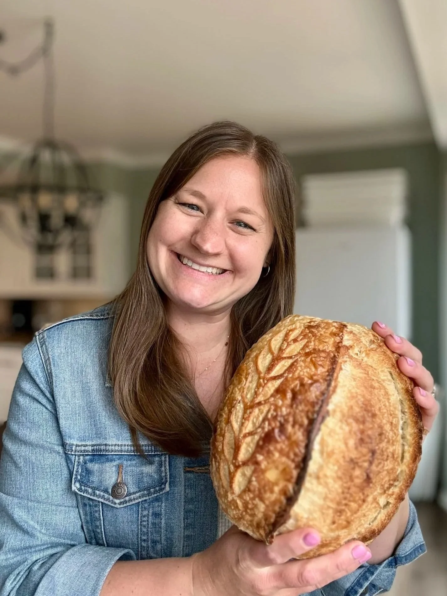 A woman with long brown hair smiling while holding a large loaf of bread.