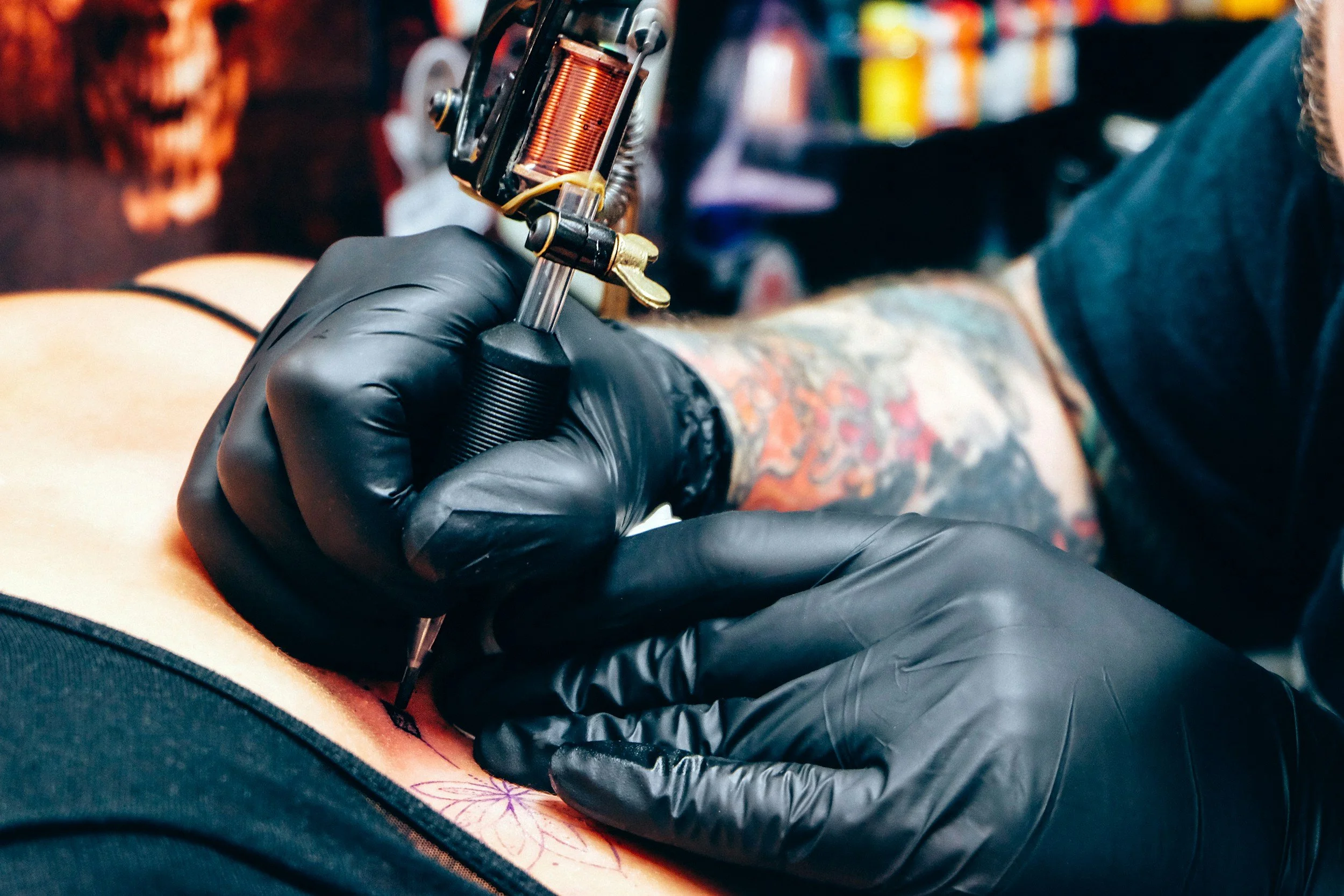A tattoo artist wearing black gloves is tattooing a colorful tattoo on a person's arm in a tattoo shop.