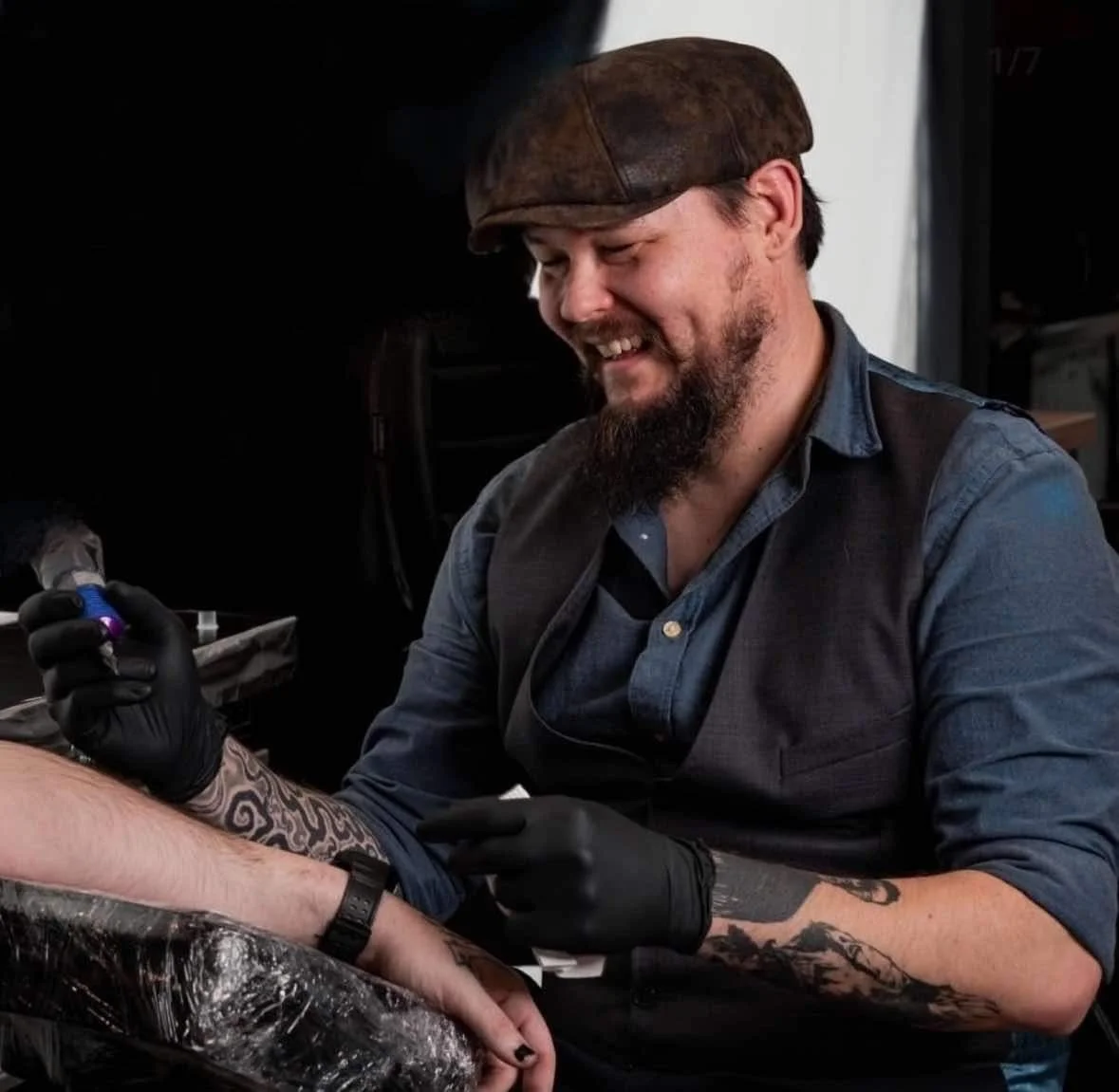 A man with a beard and tattooed arm getting a tattoo on his forearm from a tattoo artist with a beard, wearing a brown cap, gray shirt, and black gloves, inside a tattoo studio.